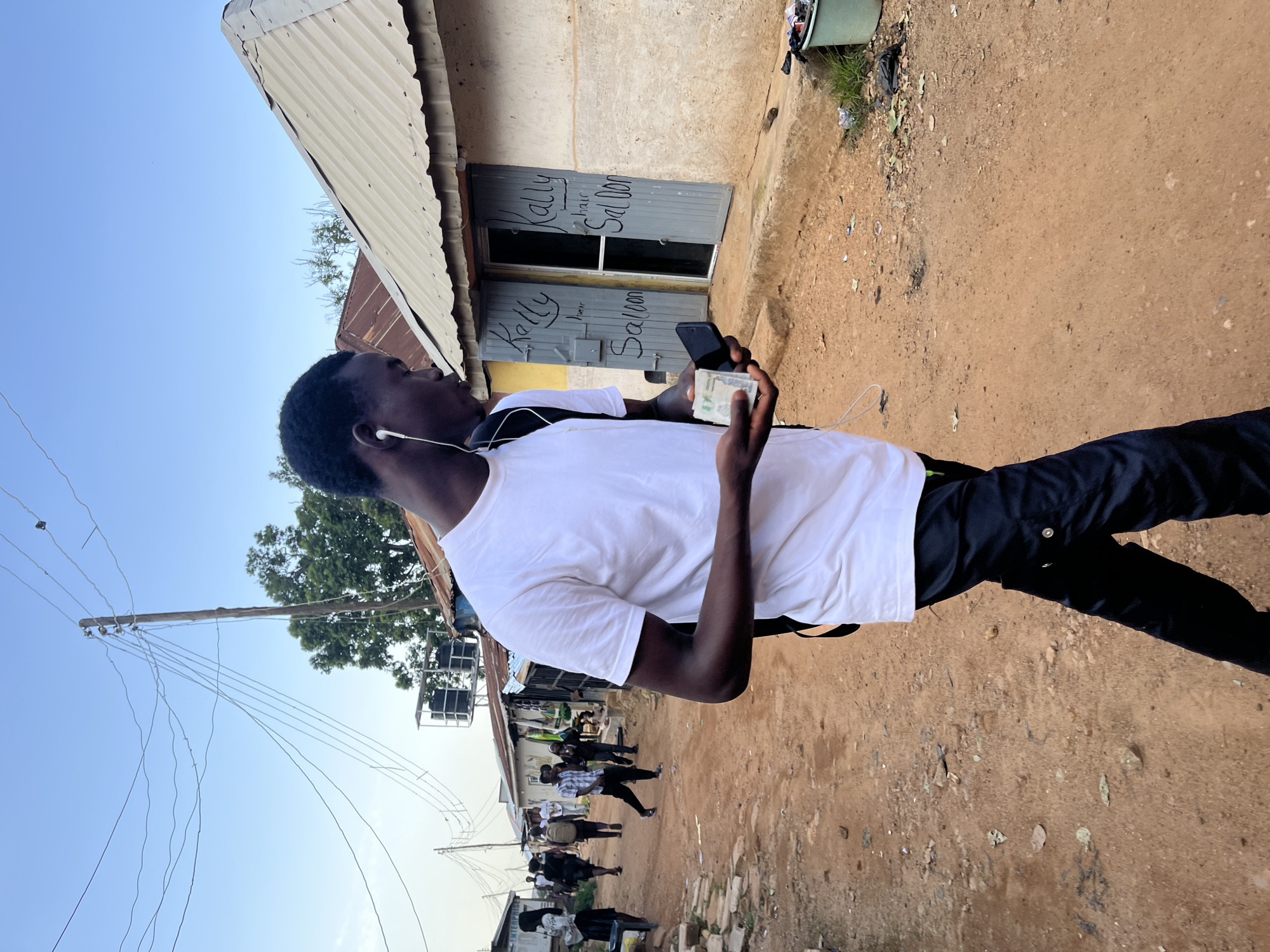 The image depicts a young Black male walking on a dusty, dirt road in a neighborhood of Jos, Nigeria. He is wearing a white t-shirt, black pants, and white earbuds. In his hands, he holds a stack of Nigerian Naira currency and a black smartphone. In the background, other individuals can be seen walking along the road, and various buildings with corrugated metal roofs line the street. One building prominently displays a sign that reads "Kally Hair Salon." The environment appears to be an informal settlement with overhead power lines and scattered debris on the ground. The lighting suggests it is either early morning or late afternoon.