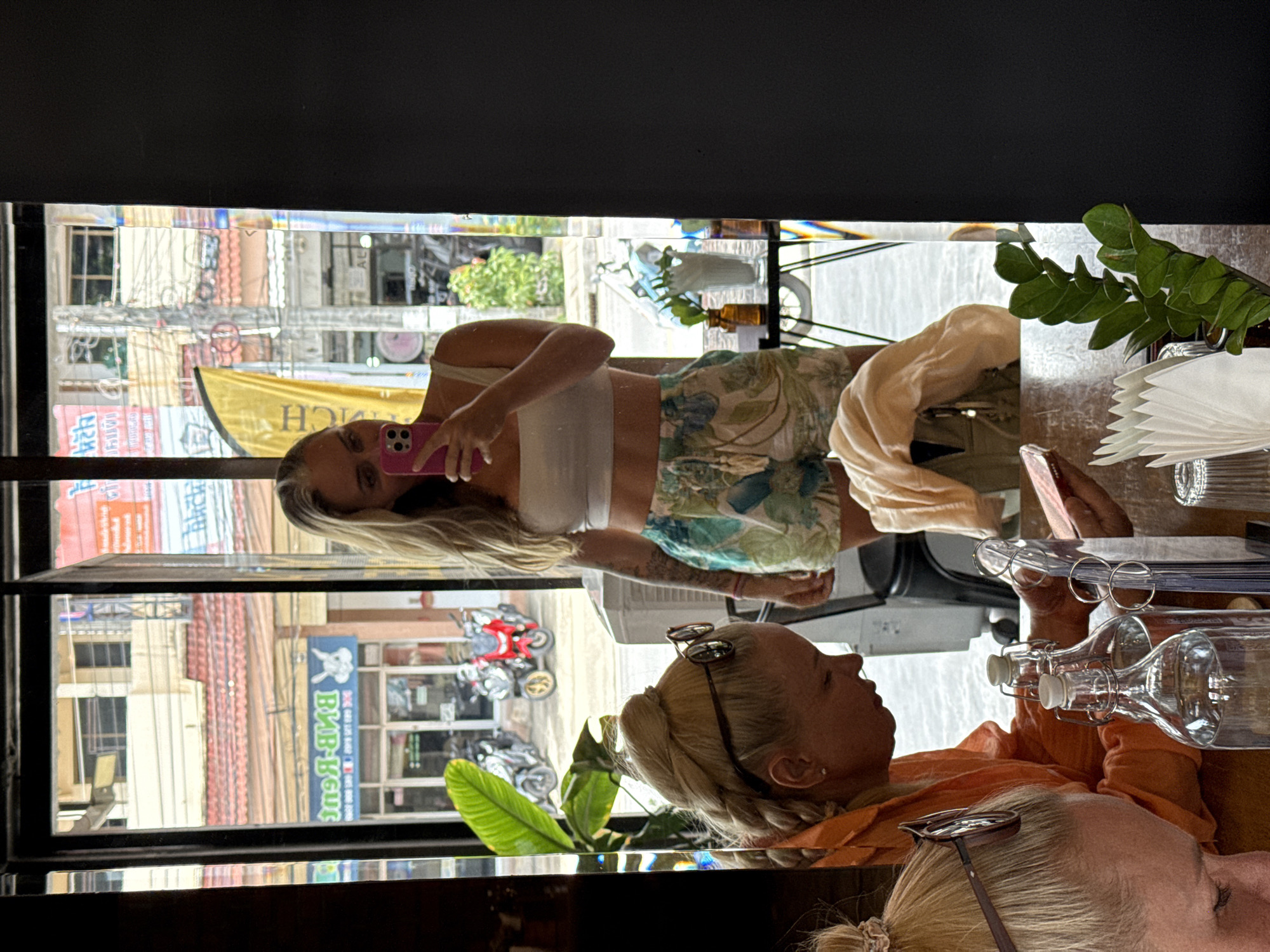 A reflection in a window shows a woman in a white crop top and floral shorts holding a pink smartphone, taking a selfie. She has blonde hair and visible tattoos on her arm. In the foreground, the reflection also shows two women at a table. The woman on the left is wearing an orange top and has her hair in a braid. The woman on the right is wearing a light-colored top and is looking down at her phone. Bottles of water and a plant are on the table. Outside the window, a street scene with buildings, motorcycles, and signs is visible, indicating the location is Ko Samui District, Thailand.