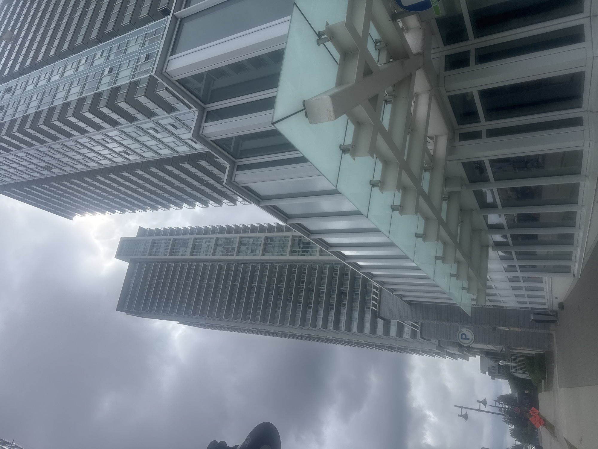The image shows a street view of two modern high-rise apartment buildings with a glass entryway in the foreground. The sky is overcast and appears to be cloudy. On the left side of the image, there is a sidewalk with streetlights and some orange construction signs. A sign with a "P" for parking is visible on the side of the building to the right of the sidewalk. The buildings are made of glass and metal. Based on the presence of street signs indicating Coquitlam, this scene is located in Coquitlam, Canada. There are no people clearly visible in the image.