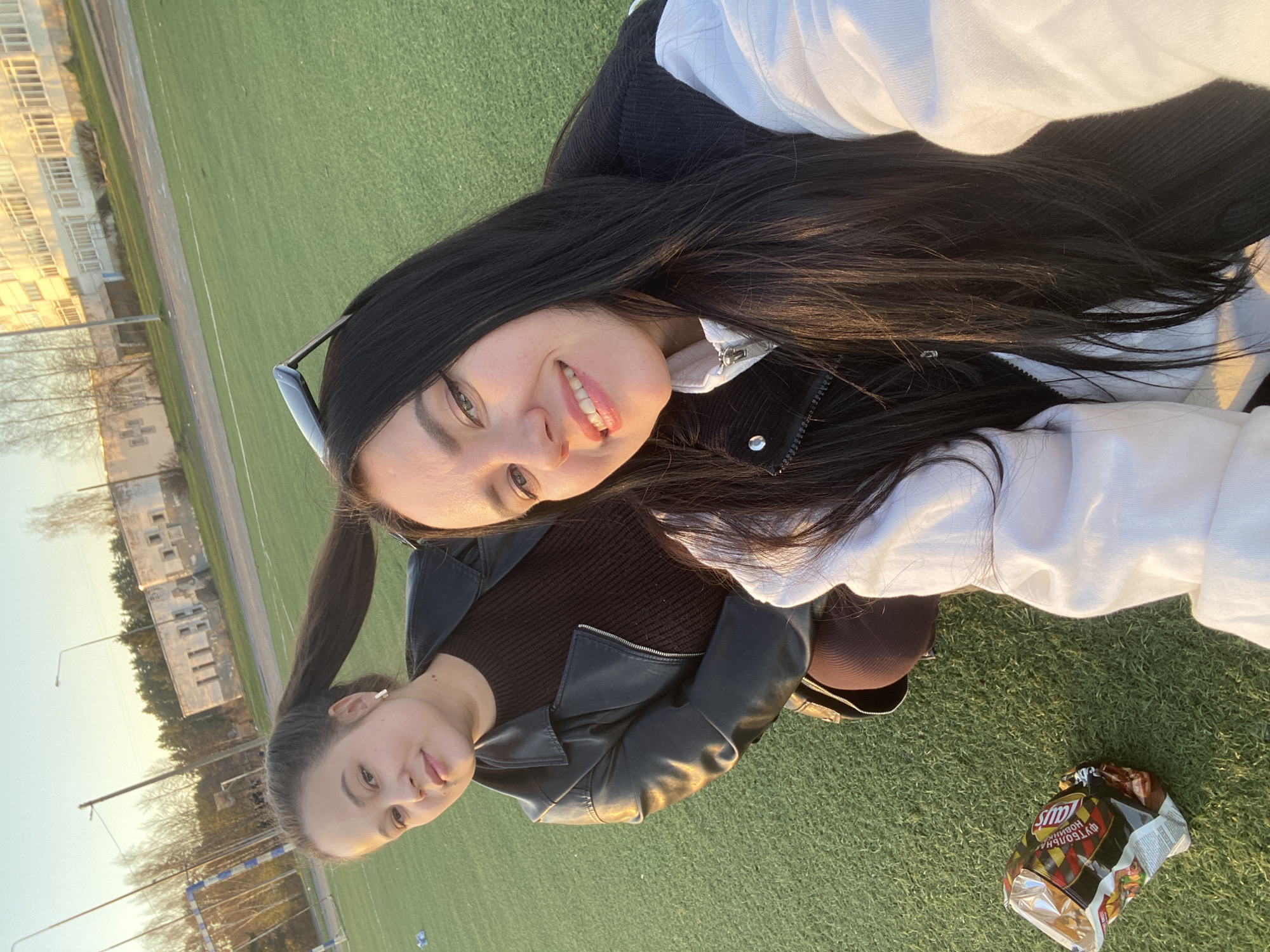 Two young women are posing for a selfie on a green artificial turf field. The woman in the foreground has long, dark hair and is wearing a white and black jacket. She is smiling. Behind her is another woman with her hair pulled back in a ponytail, wearing a dark brown shirt and a black leather jacket. In the background, there are buildings and trees, suggesting the edge of a city.
To the right of the women is an open bag of Lays chips.
Based on visual cues, the location appears to be in the city of Chaikovsky, Russia. This is determined by the lettering on the chip bag which indicates a brand widely sold in Russia.