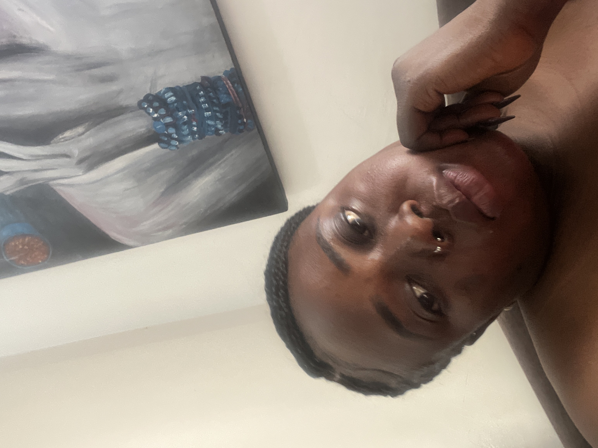 The image shows a close-up, angled shot of a woman's face. She has dark skin, braided hair, and is wearing a nose ring. Her right hand is resting on her cheek, with long, pointed fingernails. In the background, a painting hangs on a light-colored wall. The painting depicts a figure with gray fabric and a blue beaded bracelet. There are no discernible elements in the image that specifically indicate the location as Abuja, Nigeria.