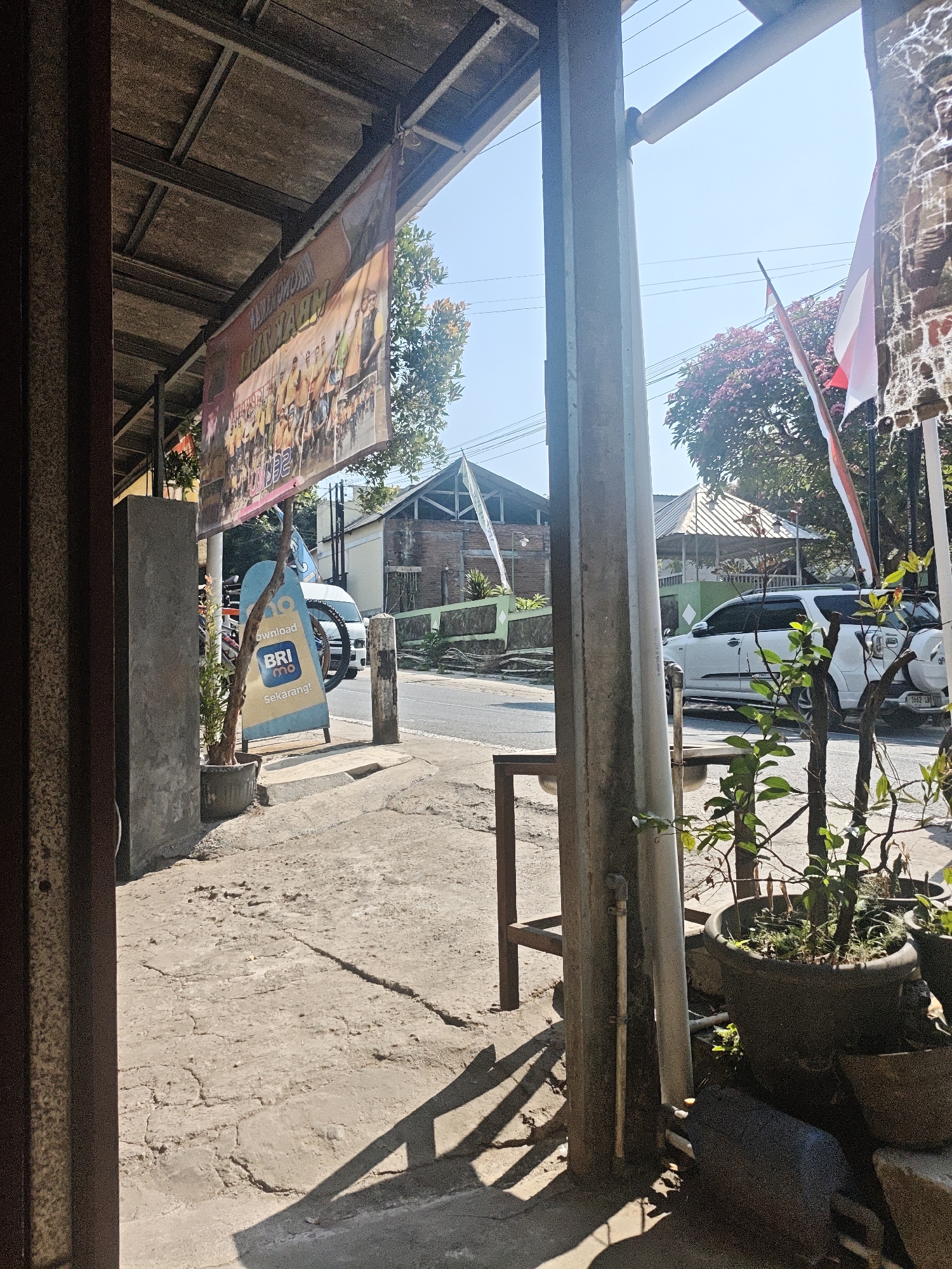 The image depicts a street scene in Lau, Indonesia, on a sunny day. In the foreground, a weathered, grey concrete pillar stands prominently, casting a long shadow across the cracked, sun-baked pavement. To the left, a dark, vertical wooden structure suggests an entrance or a part of a building. A banner with images of people, possibly participants in an event, hangs from a metal frame overhead.

A blue and yellow sign advertising "BRImo" is propped up on the sidewalk, with a potted plant beside it. Further down the street, a white SUV is parked, and a white van is visible in the background. Indonesian flags, red and white, are displayed on poles. The environment includes buildings with traditional architecture, a lush green tree with pink blossoms, and utility wires crisscrossing the clear blue sky. The overall impression is of a warm, day in a semi-urban or rural Indonesian setting.
