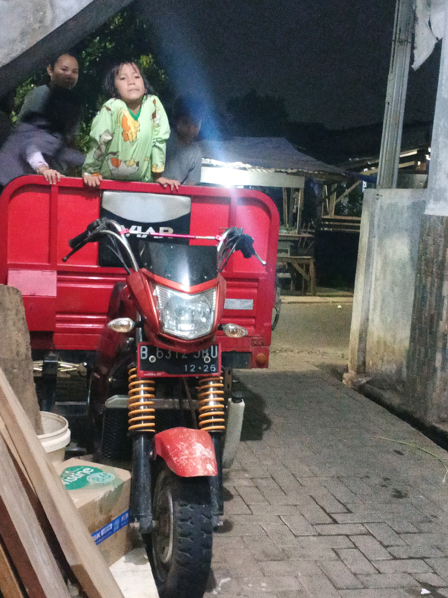 In Kadu, Indonesia, a red motorized tricycle with a cargo bed is parked on a paved surface. A young girl wearing a light green and yellow patterned pajama set is leaning over the side of the tricycle's cargo bed, looking towards the camera. She is positioned between two adults who are partially visible behind her. One adult, likely a woman, is to her left, with their hands resting on the edge of the cargo bed. Another adult is partially visible to her right. The tricycle's front is a red motorcycle with a prominent headlight and a license plate that reads "B.6312 JGU" and "12.26". The background shows a dimly lit outdoor environment with trees and what appears to be a simple structure with a corrugated roof. To the left of the tricycle, there are wooden planks and a white bucket. The lighting suggests it is evening or night.