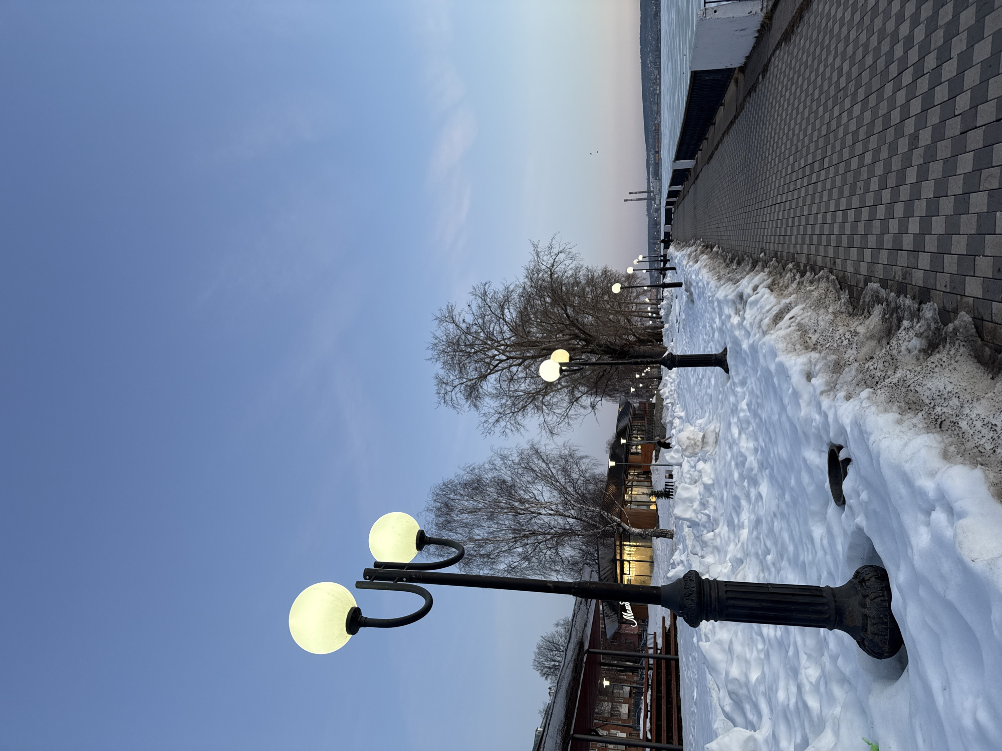 The image depicts a winter scene in Votkinsk, Russia, along a paved walkway near a body of water, possibly the Votkinsk Reservoir. The walkway is lined with decorative black lamp posts with spherical lights. Significant snow accumulation covers the ground adjacent to the walkway and partially obscures the base of the lamp posts. Bare trees stand between the walkway and nearby buildings, some of which appear to be shops or restaurants. The sky is a gradient of blue and light orange, suggesting early morning or late afternoon. In the distance, across the water, are structures that may be industrial, possibly part of a manufacturing complex. No people are clearly visible.