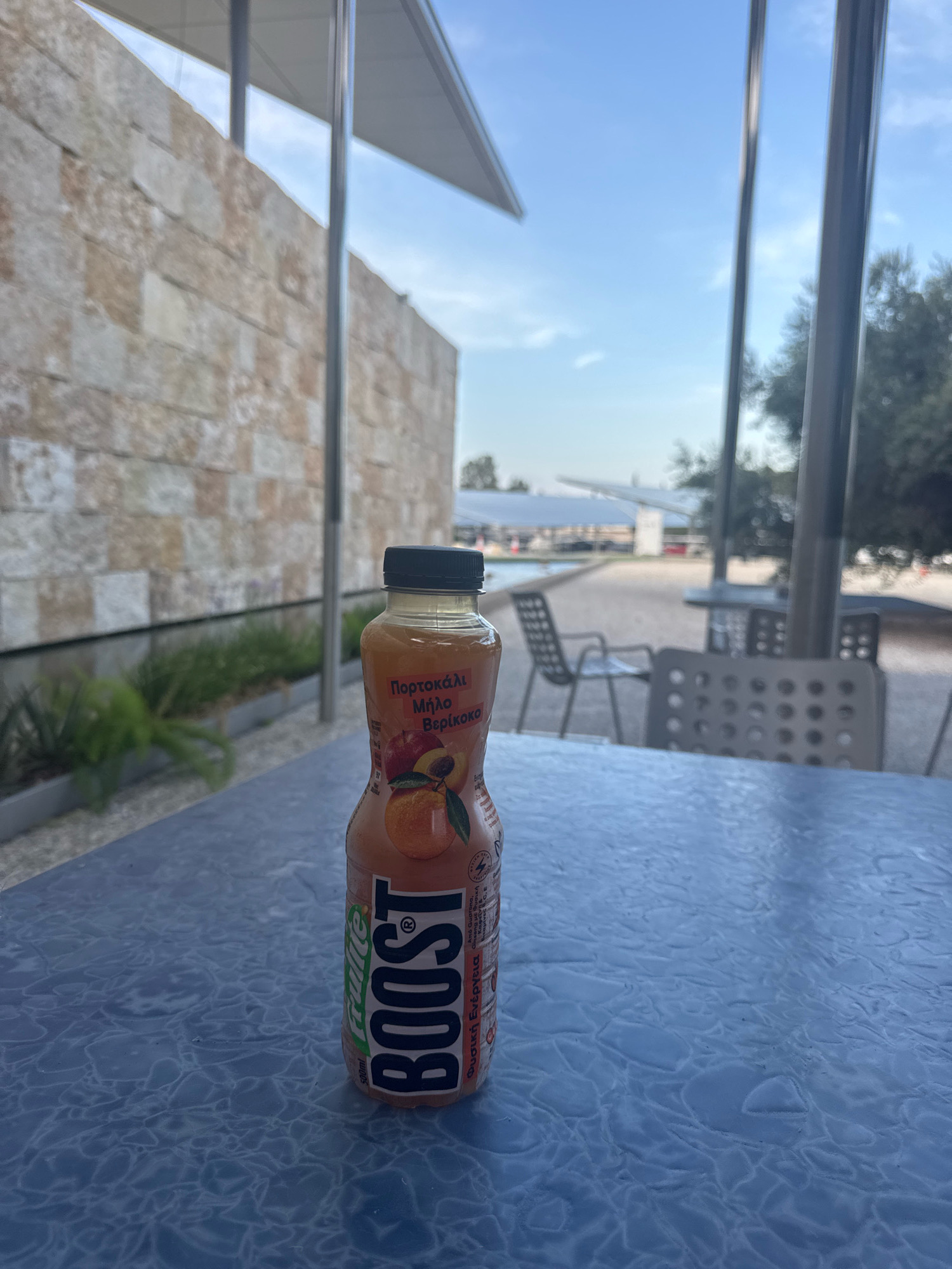 A bottle of Boost brand juice sits on a textured blue table. The bottle is partially visible on the right side of the frame. The label on the bottle features an illustration of fruit and the brand name "BOOST". In the background, a light-colored stone wall is visible, with greenery in front of it. Beyond the wall, there is an outdoor seating area with chairs and tables. The sky is blue with some scattered clouds. The scene appears to be outdoors, possibly at a cafe or restaurant in Strovolos, Cyprus.