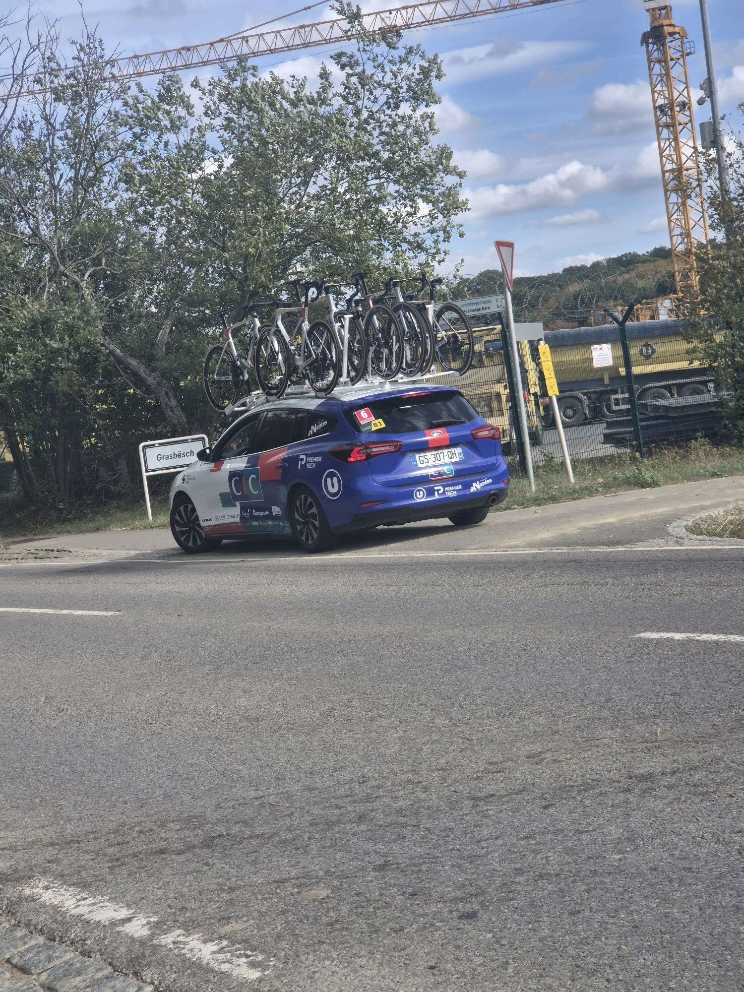 A blue car with bicycles mounted on its roof is parked on the side of a road. The car has several logos and text on its sides, including "Nantes," "PREMIER TECH," and "U." A sign to the left of the car reads "Grasbesch." In the background, there is a yellow truck, a construction crane, trees, and a fence. The sky is cloudy. The scene appears to be in Leudelange, Luxembourg, near the Grasbesch area.