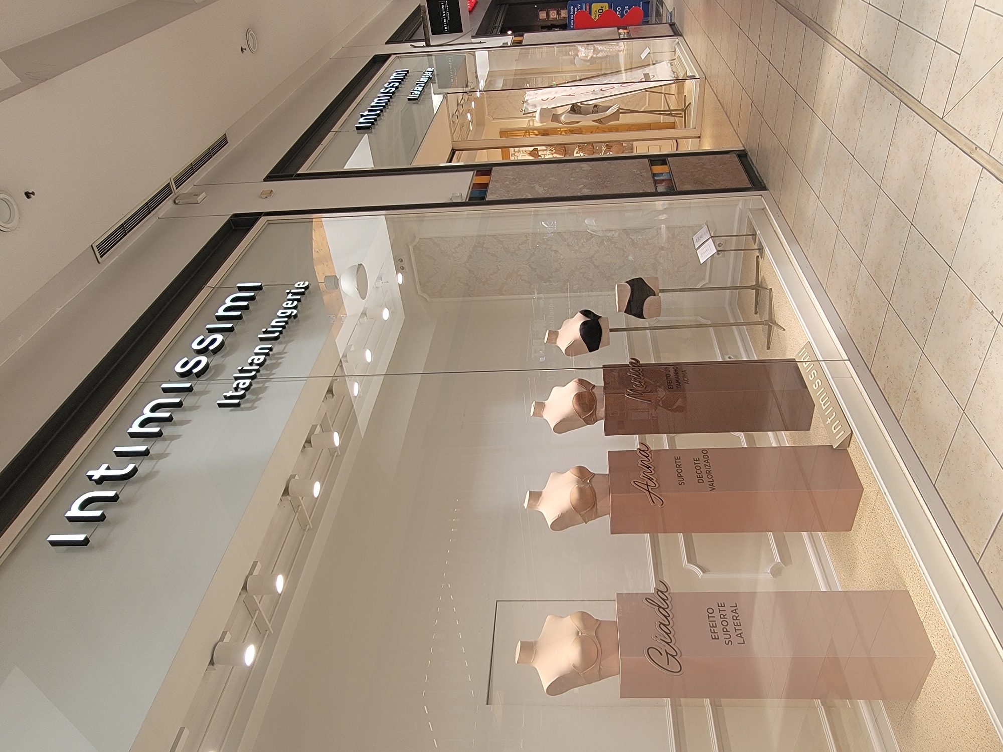 The image shows the storefront of an Intimissimi store, identified by the prominent signage. The store is located in a shopping mall, as indicated by the tiled flooring and the interior architecture. The display window features mannequins dressed in lingerie, specifically bras and underwear. One mannequin displays a black bra and black underwear, while others show bras in neutral tones. Rectangular pedestals with text in Portuguese are placed in front of the mannequins, describing product features such as "SUPORTE" (support), "DECOTE VALORIZADO" (enhanced cleavage), and "EFEITO SUPORTE LATERAL" (side support effect). The store interior is visible through the glass, showing more merchandise and display units. The exterior environment suggests an indoor shopping center, with overhead lighting and a clean, modern aesthetic. There are no visible individuals in the immediate foreground of the storefront. Based on the location provided in the prompt, this Intimissimi store is situated in Faro, Portugal.