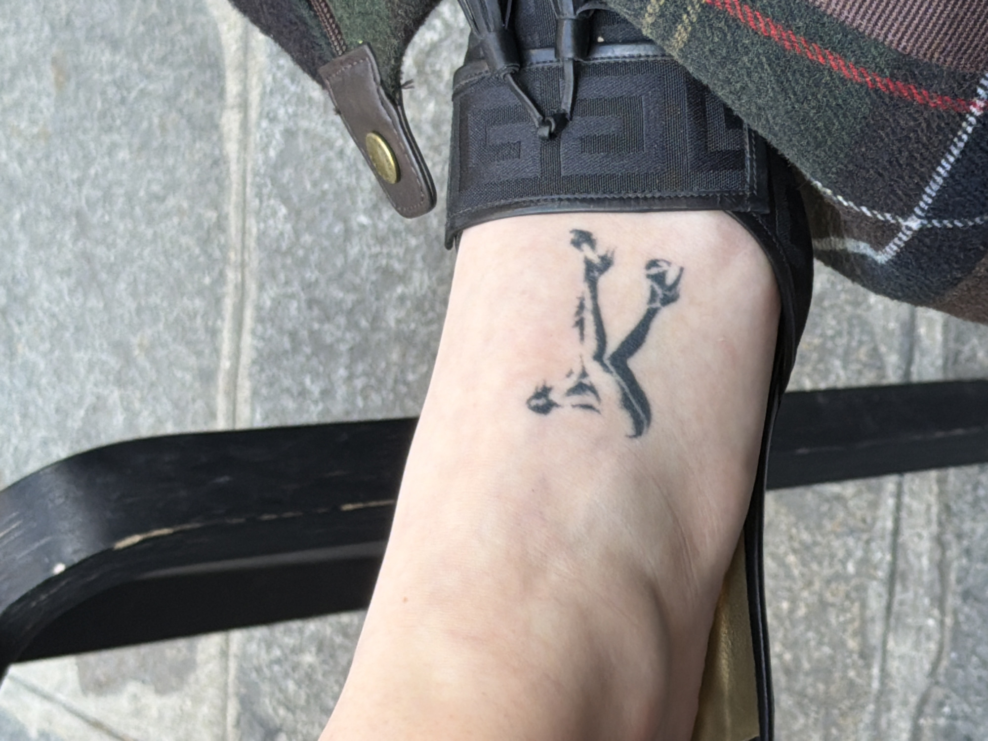 A top-down view reveals a person's left foot, adorned with a black tattoo depicting a figure, wearing a black sandal with a thick ankle strap. The person is sitting on a black bench on the ground that has a cement or concrete-like surface. Part of a patterned jacket or coat in earth tones with a red and white stripe is visible. The setting appears to be an outdoor location in Paris, France, based on the sidewalk and the bench typically found in Parisian parks or public spaces.