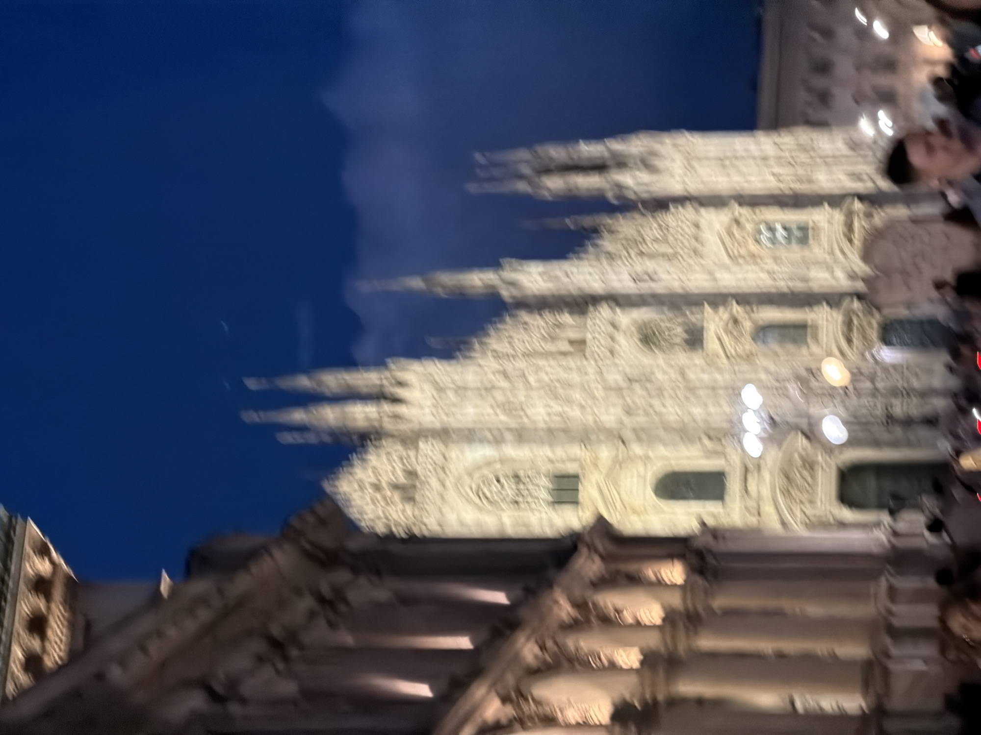 An outdoor, nighttime photograph captures a dimly lit, blurry view of the Duomo di Milano in Milan, Italy. The iconic Gothic cathedral, constructed of white marble, is illuminated by artificial lights, highlighting its intricate spires and facade. The sky above is a deep, dark blue, with a hint of clouds and a sliver of moon. To the left of the frame, a portion of a classical building with columns and arched windows is visible, also lit by warm light. In the foreground, a crowd of people are present, mostly indistinct due to motion blur, suggesting a lively atmosphere in the square. The overall impression is one of a vibrant evening scene at a famous landmark.