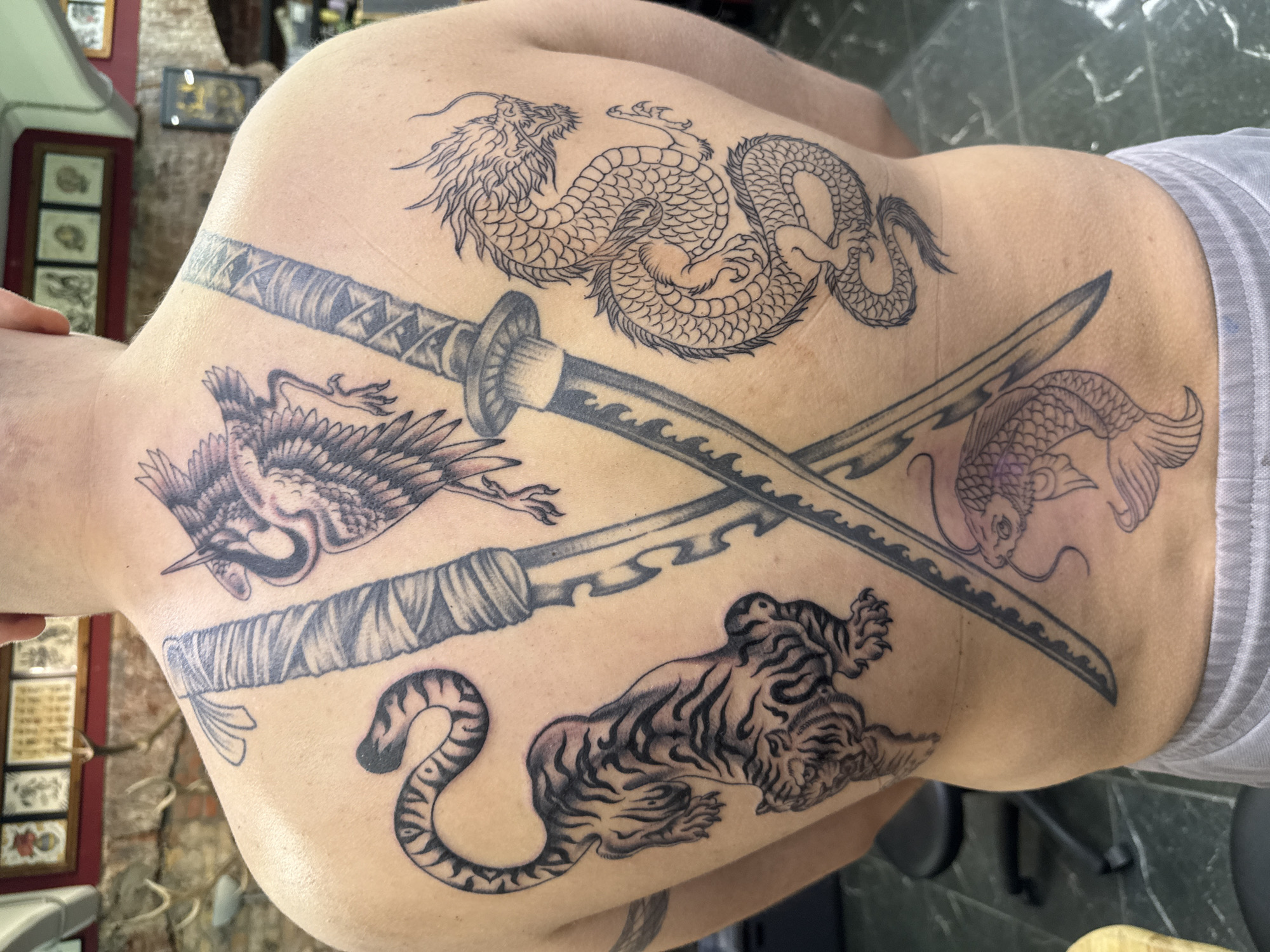 The image displays the back of an adult individual, showcasing a full array of black ink tattoos, within a setting identified as a tattoo parlor in the city of Moscow, Russia. Two Japanese-style swords (katanas) are prominently tattooed, crossed diagonally across the subject's back, with one extending from the upper left shoulder towards the lower right, and the other from the upper right shoulder towards the lower left. In the upper central back, above the intersection of the sword hilts, a detailed crane in flight is depicted. To the left of the crossed swords, on the mid-back, a crouching tiger tattoo with distinctive stripes is visible. On the right mid-back, a traditional serpentine dragon, rendered with scales and sharp claws, winds vertically. Below the intersection of the swords, on the lower back, a koi fish is tattooed, swimming upwards. The background features framed art on the upper left wall, beneath which antlers are mounted. A dark stool or chair is partially visible on the lower left, and the floor is a dark, reflective surface, possibly tiled.