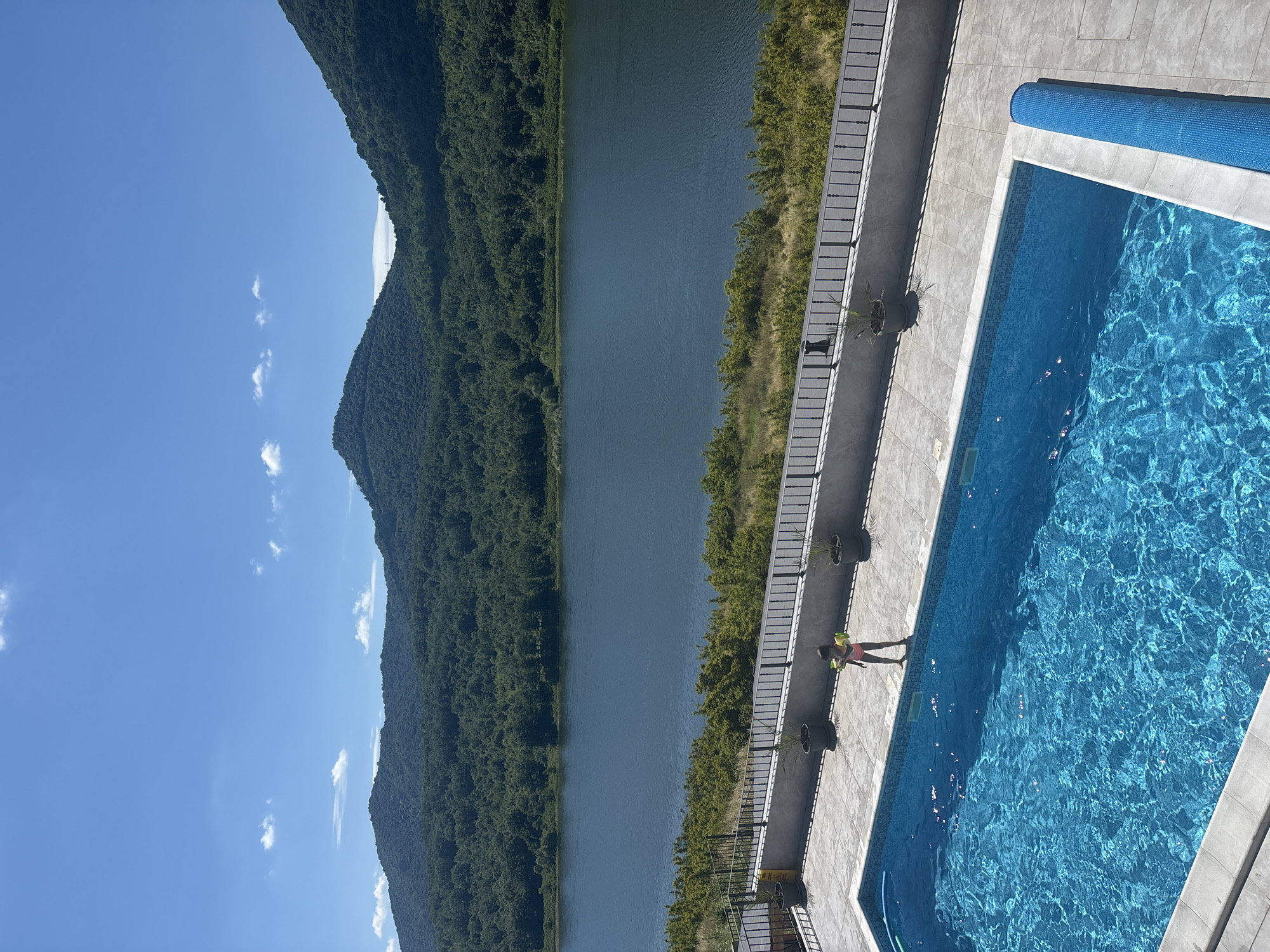 The image captures a serene outdoor scene on a sunny day. In the foreground, a rectangular swimming pool, filled with clear blue water, occupies the lower left portion of the frame. The pool is partially covered on the right side by a rolled-up blue solar cover. A young person, wearing a pink swimsuit, stands on the tiled edge of the pool, holding yellow and green objects in their hands, possibly toys.
Beyond the pool area, a stone patio is visible, bordered by a black metal fence. Several potted plants are placed along the patio. The middle ground features a wide, calm body of water, likely a lake or reservoir, reflecting the bright blue sky. The water is bordered by a strip of green vegetation and trees.
In the background, rolling hills densely covered with lush green trees dominate the landscape. The sky above is a clear, vibrant blue, with a few wispy white clouds scattered across it. The overall impression is one of a tranquil, picturesque location, likely a resort or private residence, situated amidst natural beauty.