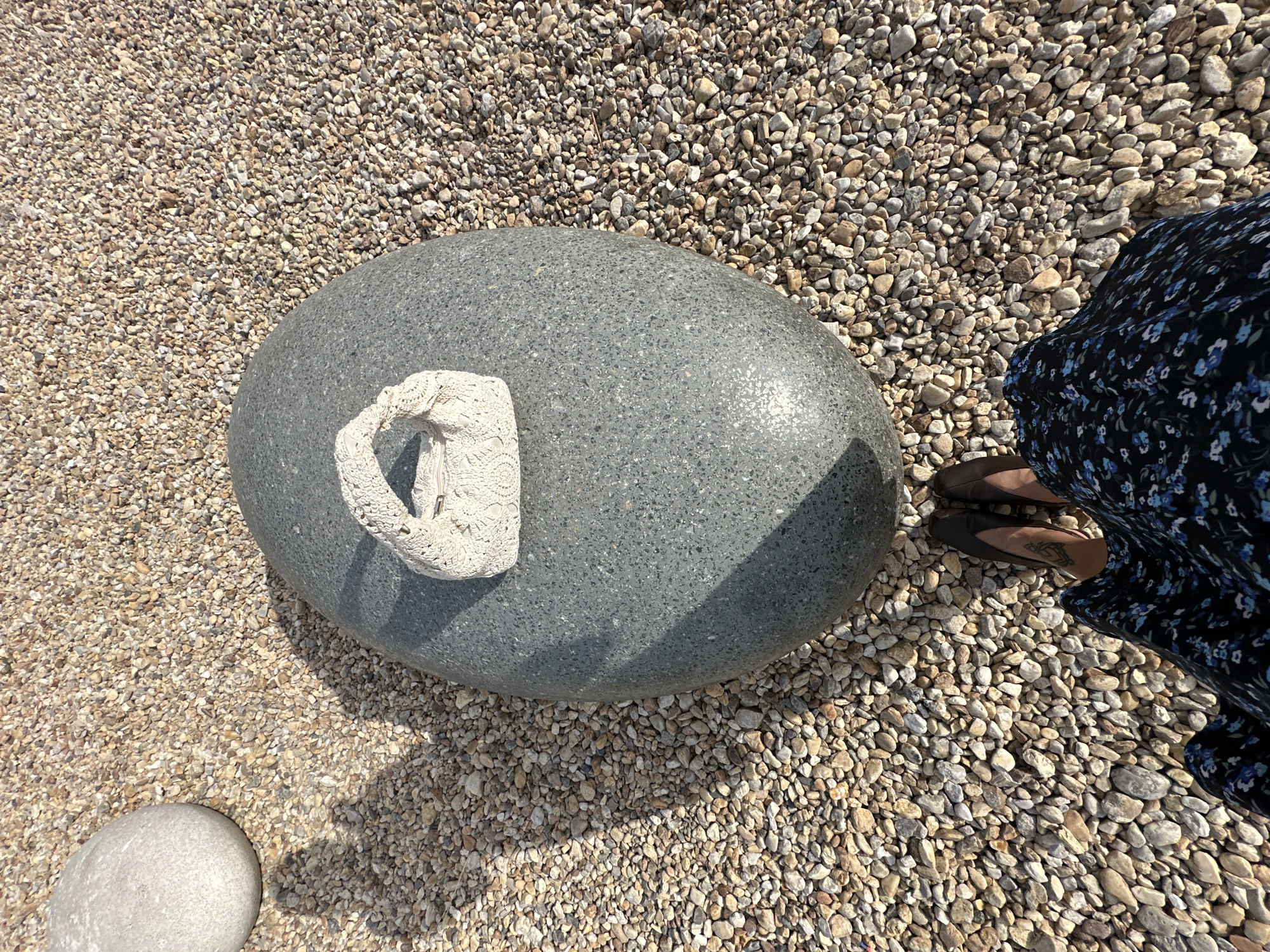 A close-up, overhead shot shows a person's legs and feet standing on a gravel-covered surface. The person is wearing a dark blue floral print dress and brown slip-on shoes. A tattoo is visible on the person's left ankle.
In the center of the image is a large, smooth, grey, oval-shaped stone. Resting on top of this stone is a small, white, crocheted object with an intricate, lace-like pattern. Another smaller, lighter-colored stone is visible in the lower left corner of the frame. The ground is covered in a variety of small, tan, grey, and white pebbles and stones.
The lighting suggests it is daytime, with a clear shadow cast by the large grey stone, indicating bright sunlight. The scene appears to be outdoors, likely a beach or a decorative garden area. No text is visible in the image. The overall mood is calm and serene. The location is Yuzhnouralsk, Russia.