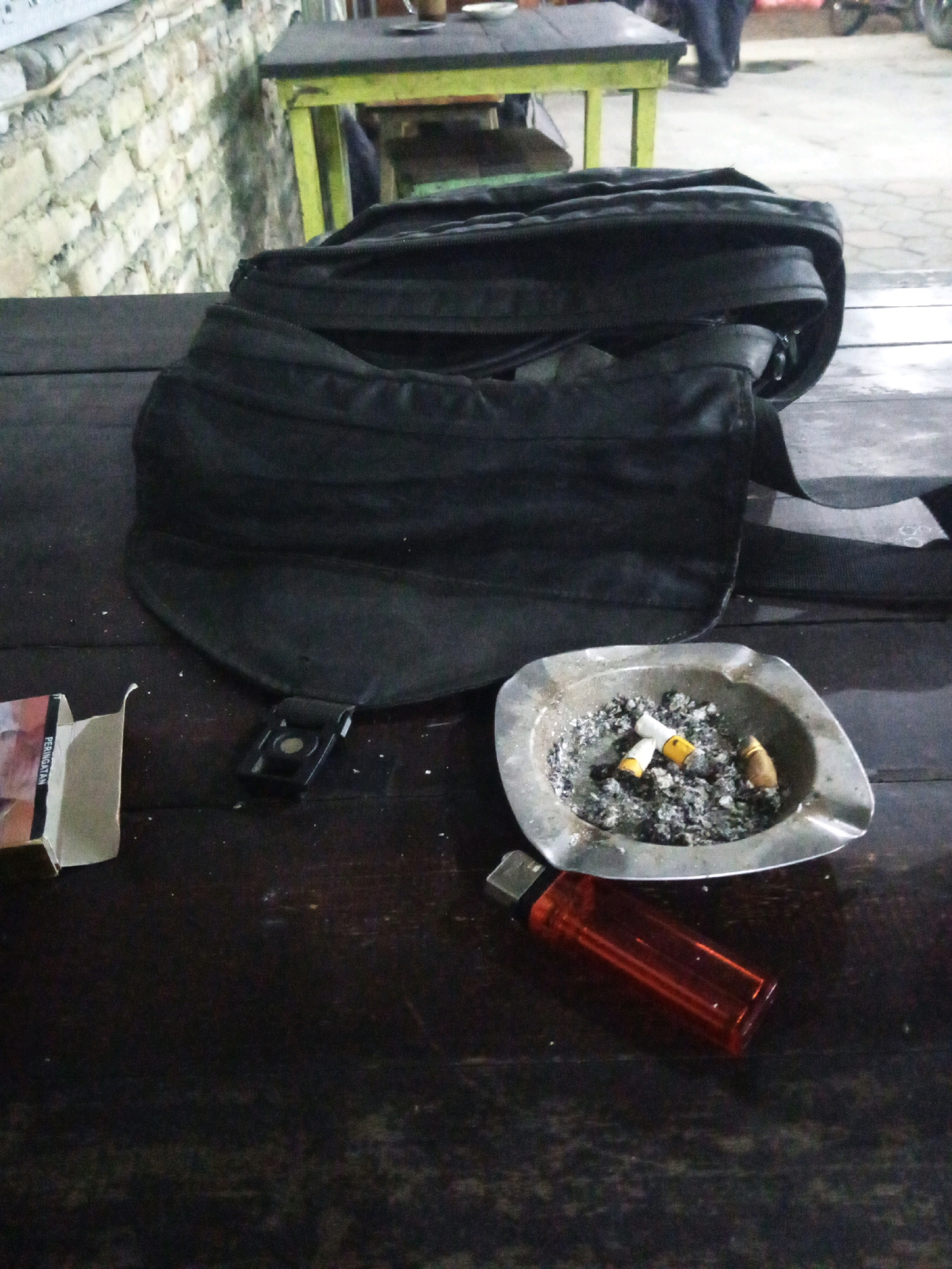 The image depicts a close-up scene on a dark wooden table, likely in an outdoor setting given the brick wall and suggestion of a paved area in the background. The primary focus is a black backpack placed centrally on the table. To the left of the backpack is a partially opened pack of cigarettes, with the warning label "PERINGATAN" visible. Adjacent to the cigarette pack is a small black clip. In front of the backpack, towards the right side of the table, is a metal ashtray filled with cigarette butts and ash. A red plastic lighter lies in front of the ashtray, oriented diagonally. The overall scene suggests a moment of pause or rest, possibly in a public area like a roadside stall or cafe. The environment appears to be dimly lit, with the brick wall indicating a rustic or older building construction. The exact location in Gayaman, Indonesia, is not discernible from the visual elements alone, but the presence of the Indonesian warning label on the cigarette pack points to the country.