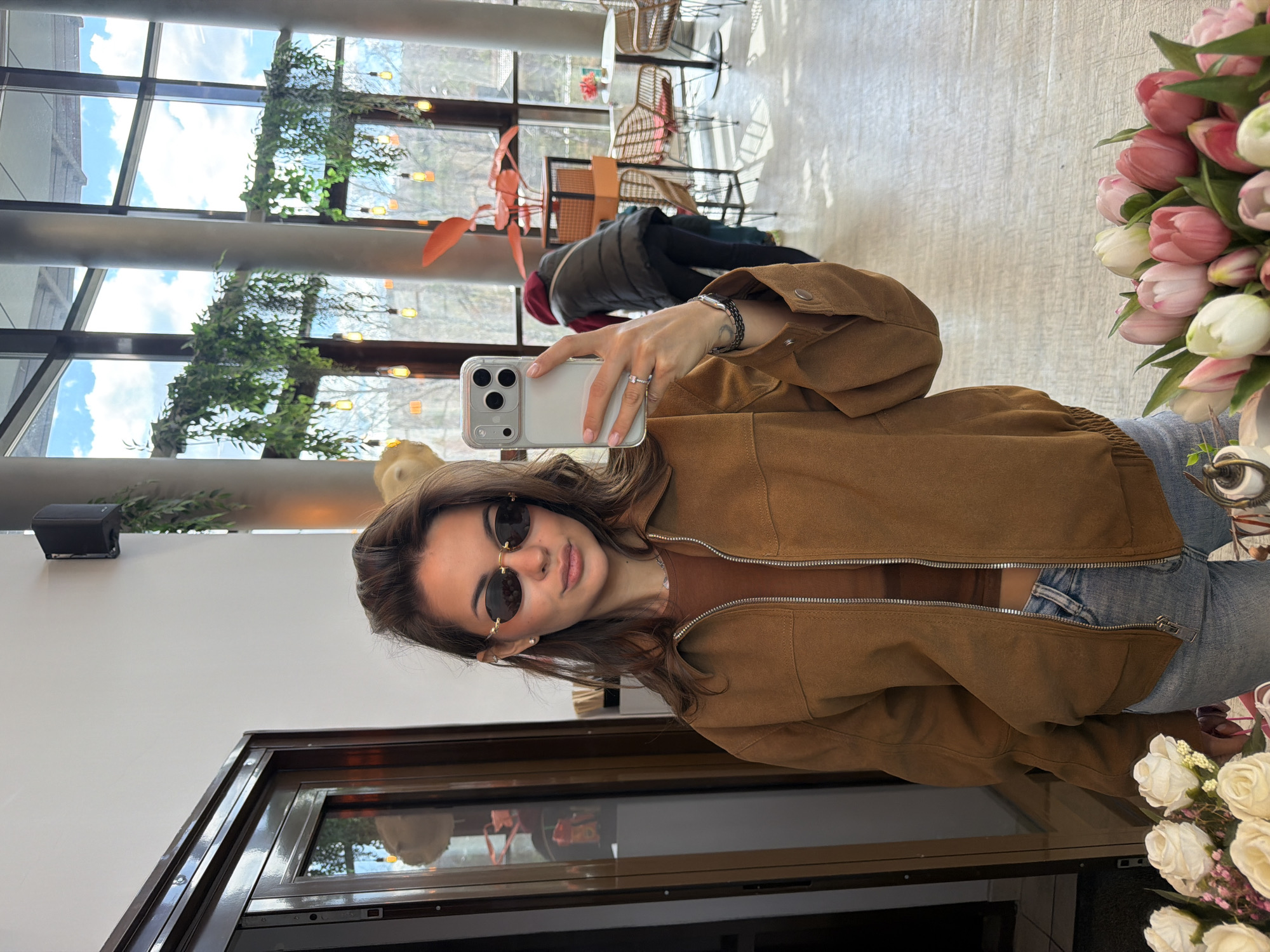 A young woman is taking a selfie in a brightly lit interior setting. She has brown hair and wears round sunglasses, a brown jacket, and jeans. She holds a smartphone in her right hand. The background features large windows with greenery visible outside. Tables and chairs are arranged within the space. There are flowers nearby, and a reflection in a glass surface adds to the scene. The setting appears to be a cafe or restaurant with modern décor. Although the location in the city of Krasnoyarsk, Russia cannot be verified forensically from the image alone, the setting suggests a public space in an urban environment.