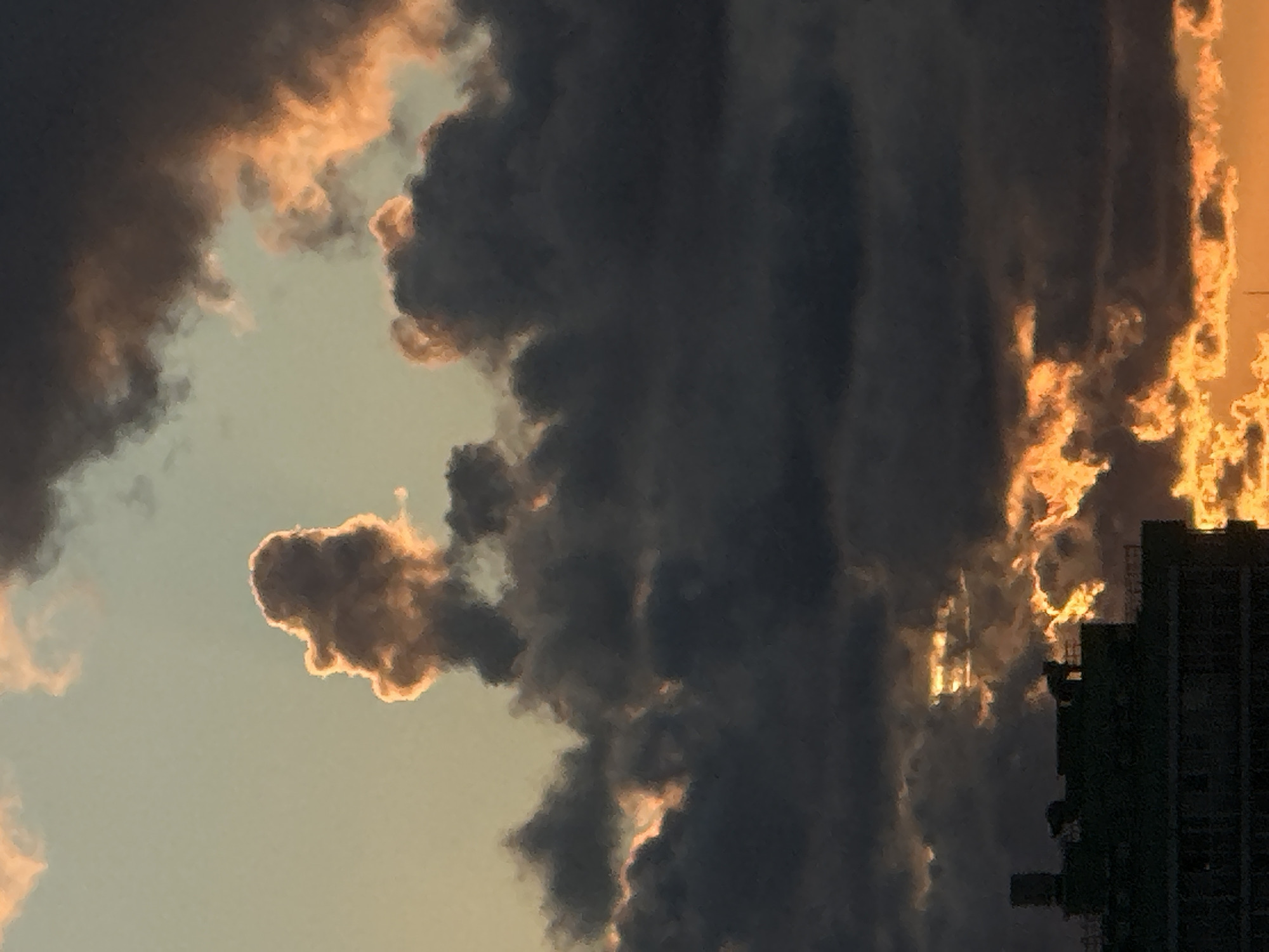 A dramatic sunset is depicted in the sky above the city of Dnp Ryzhiki, Russia. Dark, heavy clouds dominate the foreground, interspersed with vibrant streaks of orange and yellow light. In the distance, the silhouette of a building, possibly an apartment complex or industrial structure, is visible against the fiery sky. The top portion of the image shows a lighter sky with a few wisps of illuminated clouds.
