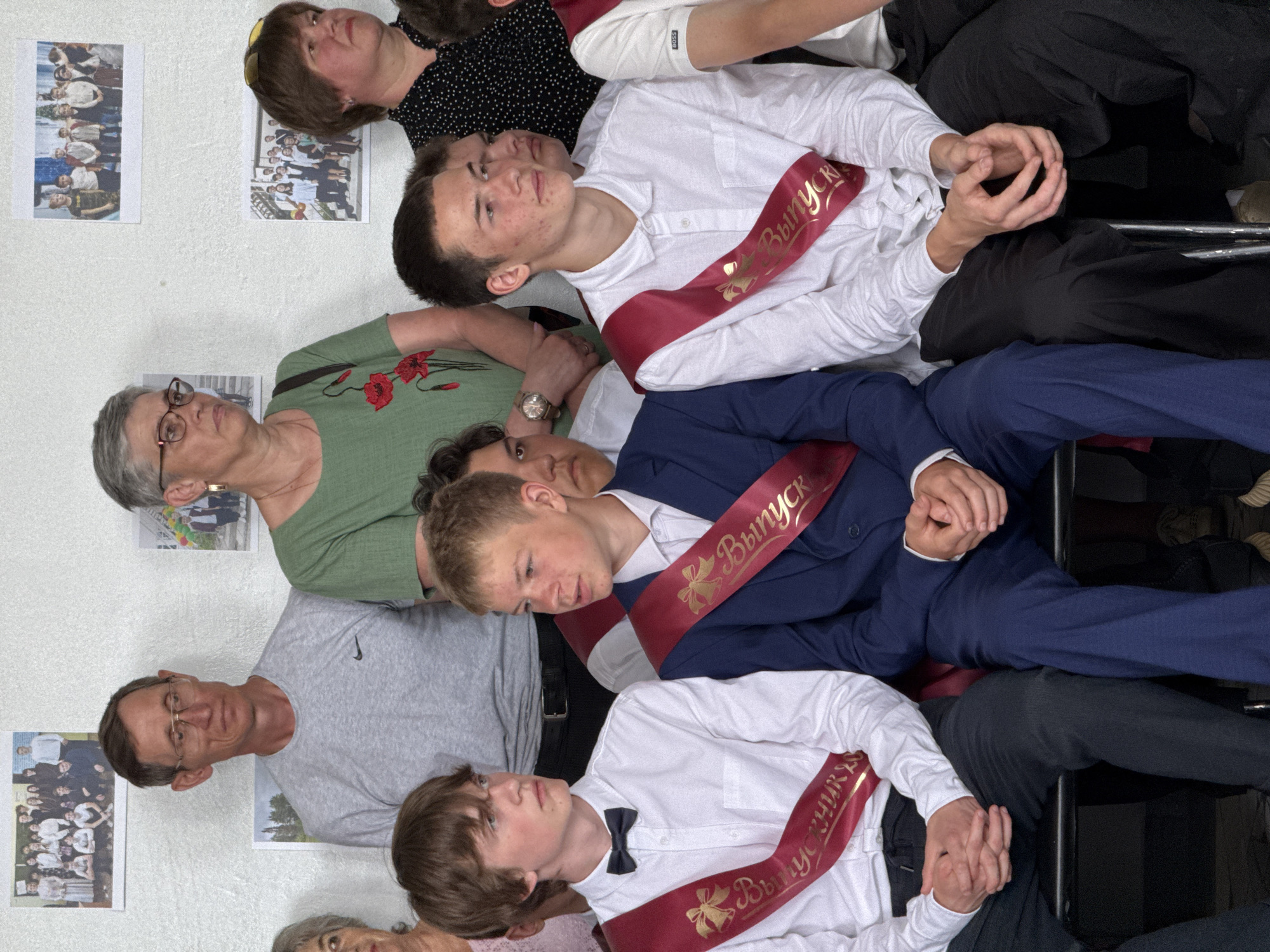 A group of people, including three young men wearing maroon sashes with gold Cyrillic lettering, are seated closely together, likely posing for a group photograph. The sashes suggest a graduation or similar celebratory event. The men are dressed in formal or semi-formal attire, with one wearing a blue suit and another a bow tie. Some adults are also present in the scene, positioned among the young men. The background features a wall with framed photographs or artwork hanging on it. The location appears to be indoors.