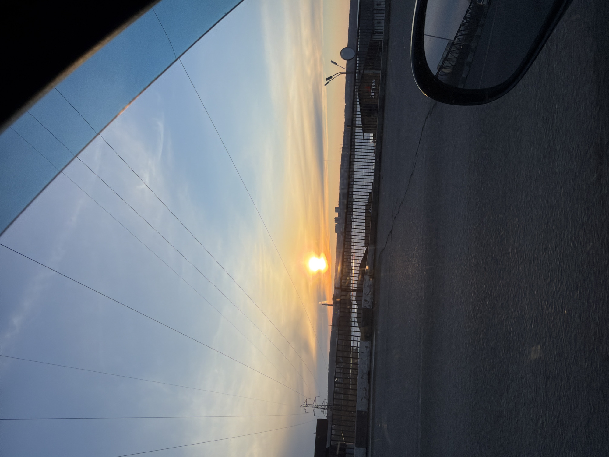 The image shows a view from inside a car, likely taken while driving. The sun is setting over the city, visible in the distance. A bridge with railings and streetlights is visible through the window. Power lines are strung across the sky. A side-view mirror of the car is also visible.
Given the visual cues and the general style of the setting, it is possible that the location is in Izhevsk, Russia, as power lines, streetlights, and urban features are common in urban areas. It appears to be evening. There are no visible people in the image. The main subjects are the sunset, the cityscape, the bridge, and the car interior.