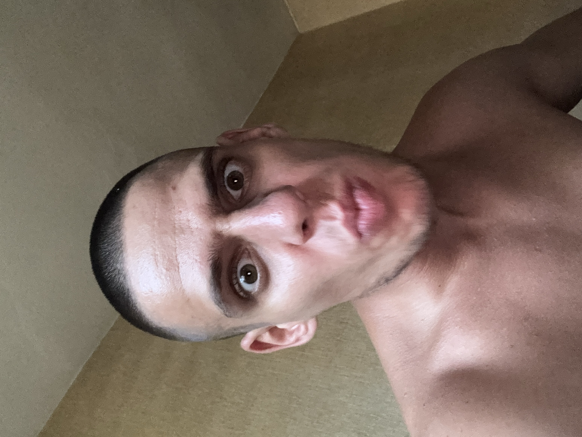 A man, with a buzzed haircut and wide, surprised eyes, makes a fish-face at the camera. He is shirtless, showing his bare chest and shoulders. The background is a textured, light brown wall, with a hint of a ceiling corner visible at the top. The man's skin appears slightly flushed, and there is a subtle stubble on his chin. The lighting is soft, suggesting an indoor setting, possibly a bedroom. The man's pose and expression suggest a playful or humorous interaction.