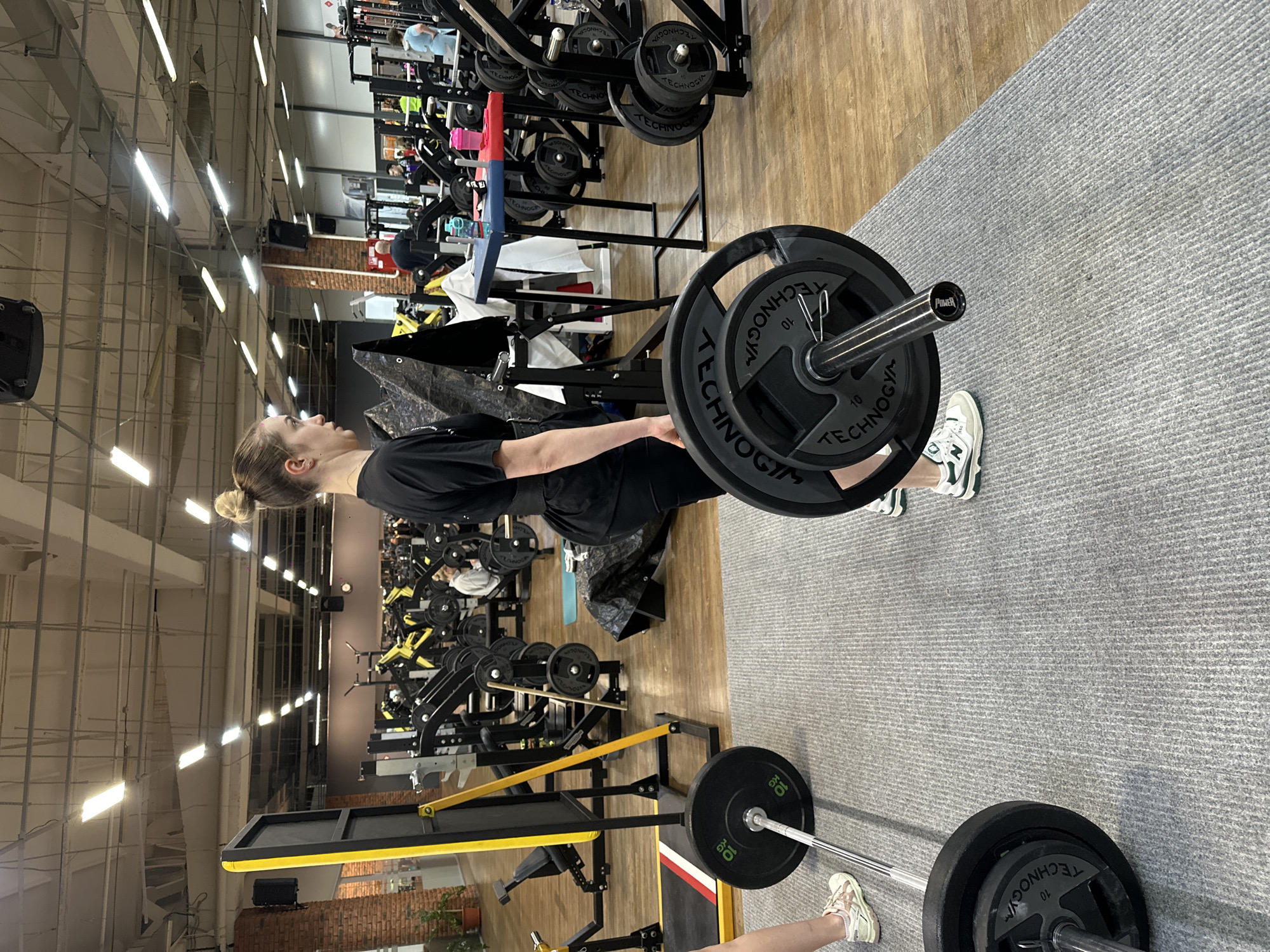 A young woman is captured in a gym in Chaikovsky, Russia, engaged in a weightlifting exercise, likely a deadlift. She is viewed from the side, wearing a black t-shirt and black shorts, with her hair pulled back in a bun. Her hands are gripping a barbell, which is loaded with two 10kg "TECHNOGYM" weight plates on each side. The barbell sleeve also displays a "POWER" logo. Her right foot, wearing a white and green "NEW BALANCE" sneaker, is visible near the barbell.
The gym features a floor divided into a wooden section and a light grey, ribbed carpeted area where the woman stands. In the background, a variety of gym equipment is present, including weight racks, other barbells, benches, and various exercise machines. Another person's legs and feet, wearing light-colored sneakers, are visible in the background near a different barbell setup.
The gym environment includes high ceilings with exposed metal structures and numerous bright fluorescent lights, indicating an indoor setting, likely during the day or evening. A section of a brick wall and a small potted plant can be seen in the midground. The overall scene depicts a focused individual participating in a strength training session within a well-equipped fitness facility.