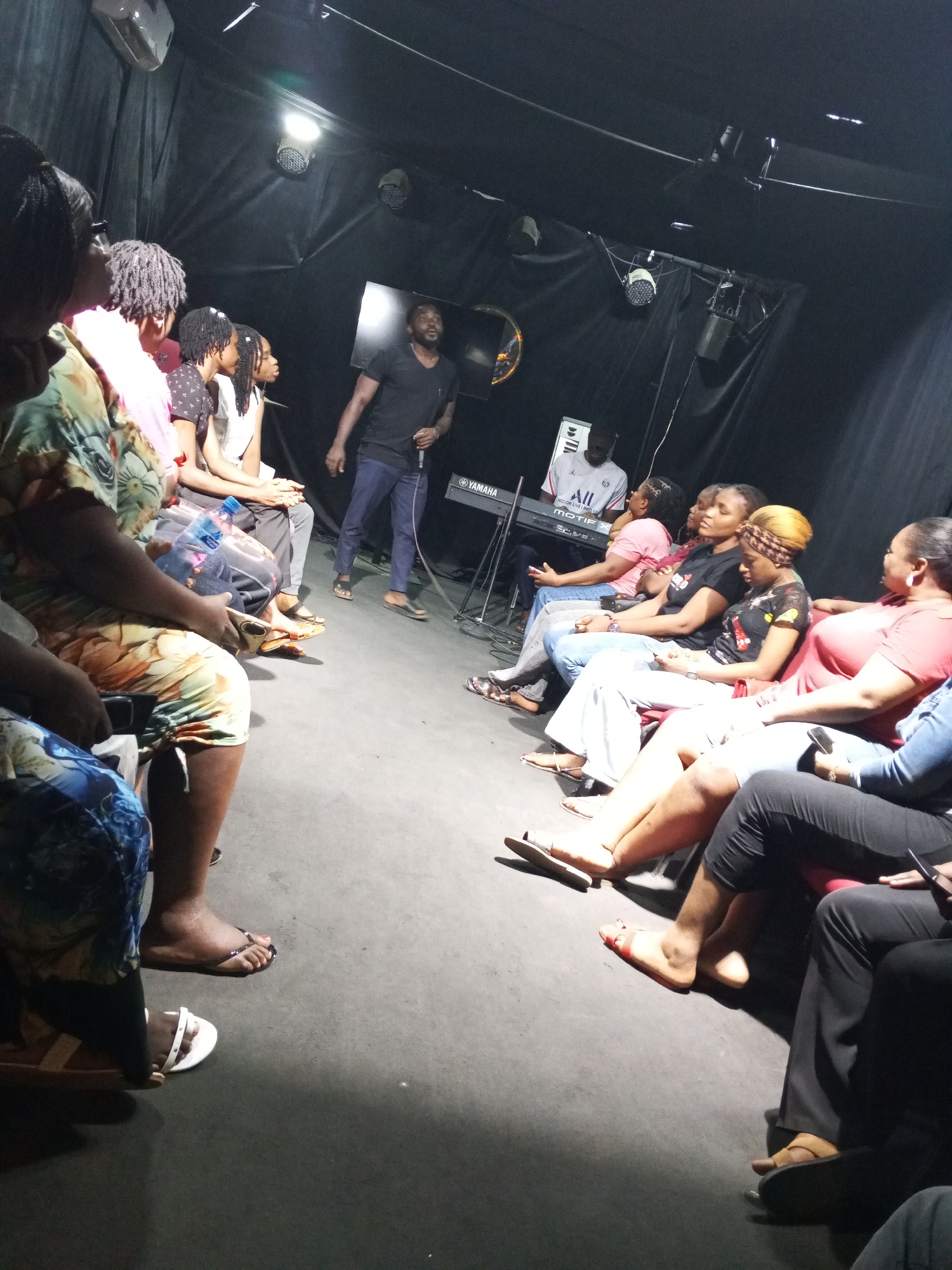 The image depicts an indoor gathering, likely in Abuja, Nigeria, given the context. The space appears to be a small, dimly lit room with a dark backdrop, possibly a stage or performance area. Approximately 15 people are visible, arranged in two facing rows, suggesting a meeting or presentation setting. One man stands facing the group, holding a microphone, indicating he is the speaker or presenter. Another man is seated behind a Yamaha Motif synthesizer. The attendees are casually dressed, and many are wearing flip-flops. There are stage lights and what appears to be a video screen in the background. The overall impression is that of a casual and informal event.