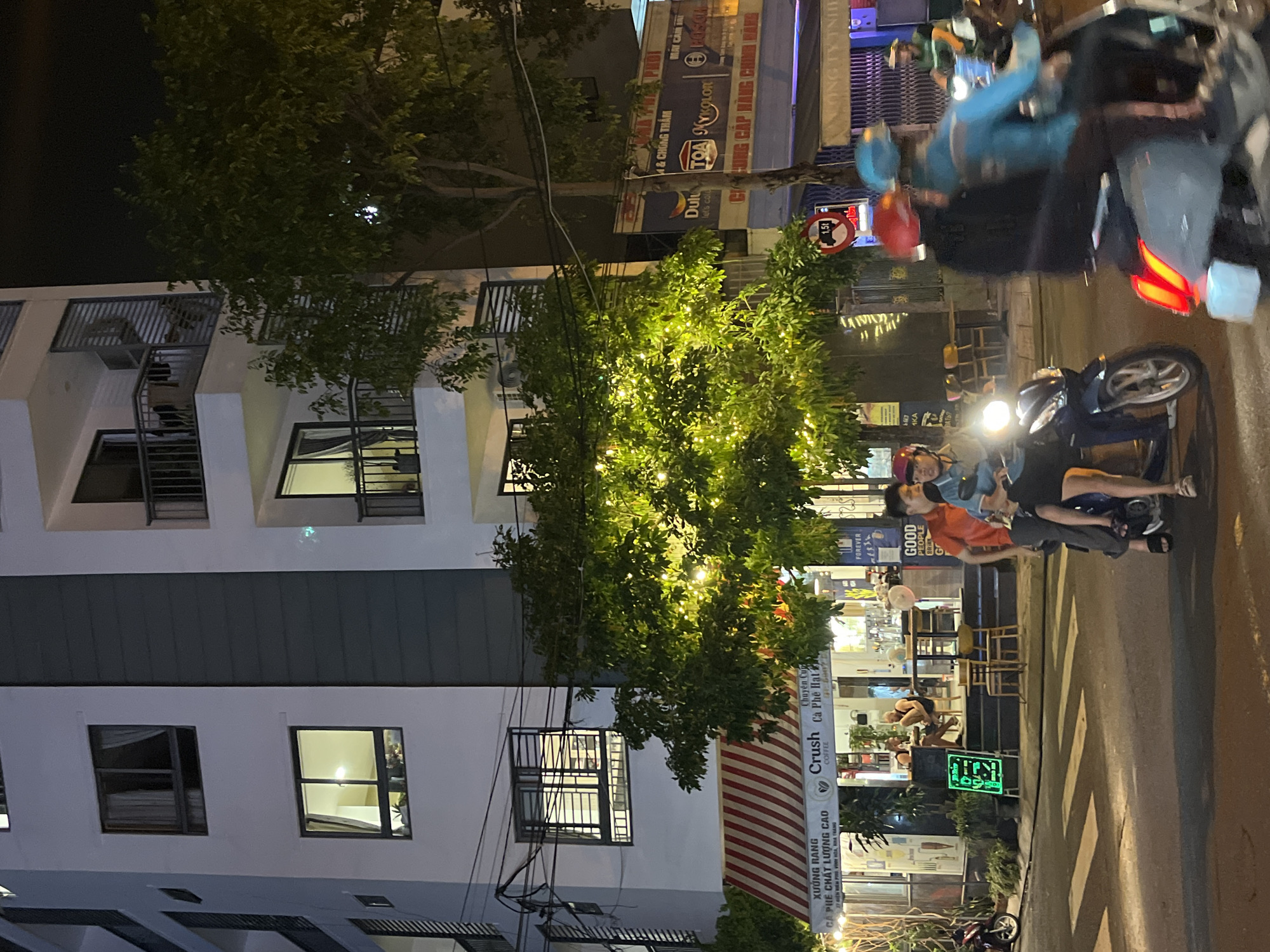 The image depicts a nighttime street scene in Bac Nha Trang Ward, Vietnam. A tall, narrow apartment building is on the left side, with lit windows indicating occupancy. A tree decorated with small lights is positioned between the building and the street. In the background, there are illuminated commercial signs in Vietnamese, suggesting a business district. A "Crush Coffee" sign is partially visible. Several people are riding a motorbike. The street is marked with zebra crossing lines.