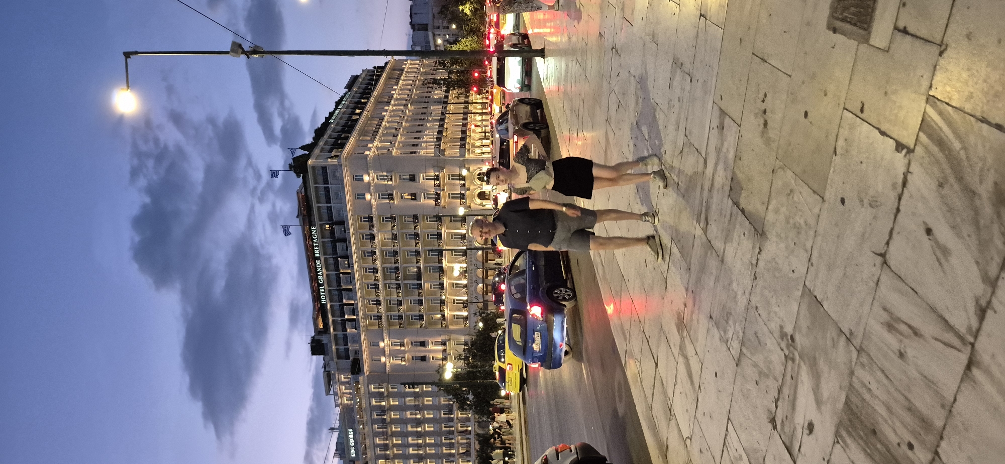 The image captures a street scene in Athens, Greece, at dusk, with the iconic Hotel Grande Bretagne as the dominant backdrop. The hotel, illuminated by warm lights, is a grand, multi-story building featuring classical architecture, balconies, and Greek flags atop its roof. In the foreground, two individuals, a man and a woman, are walking on a marble-tiled sidewalk. The man is wearing a dark polo shirt and grey shorts, while the woman is in a patterned short-sleeved top and a black skirt. They appear to be walking in the same direction.
The street in front of them is busy with traffic. Several cars are visible, including a yellow taxi and a blue hatchback. The streetlights and car headlights cast reflections on the wet pavement, suggesting recent rain or a damp surface. The sky is a deep twilight blue, with scattered clouds. Other buildings and streetlights are visible in the background, contributing to the urban atmosphere. The scene conveys a sense of a city transitioning from day to night, with people enjoying a walk and traffic continuing its flow.