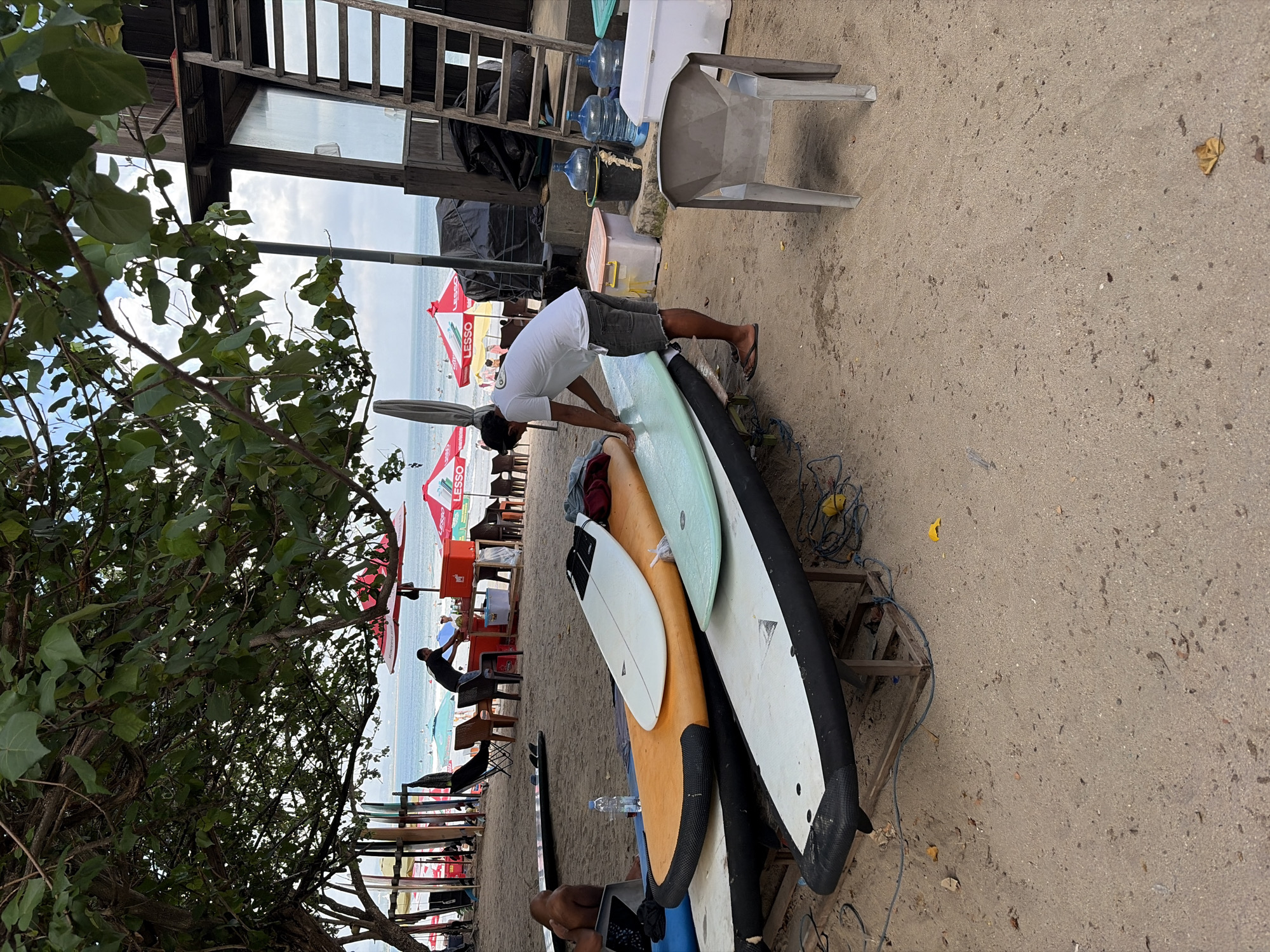 The image depicts a beach scene in Kuta, Indonesia, likely near a surf rental or school. In the foreground, a man in a white t-shirt and dark shorts is standing over several surfboards, appearing to be working on one of them. Three surfboards are stacked on a wooden rack, and another is laid out in front of them. The surfboards are various colors, including orange, white, and light green. The ground is covered in sand, with some scattered debris. In the midground, there are beach umbrellas, chairs, and a small building with a wooden ladder leading up to it. Beyond that, the ocean is visible with people in the water. The overall impression is of a busy, casual beach environment.