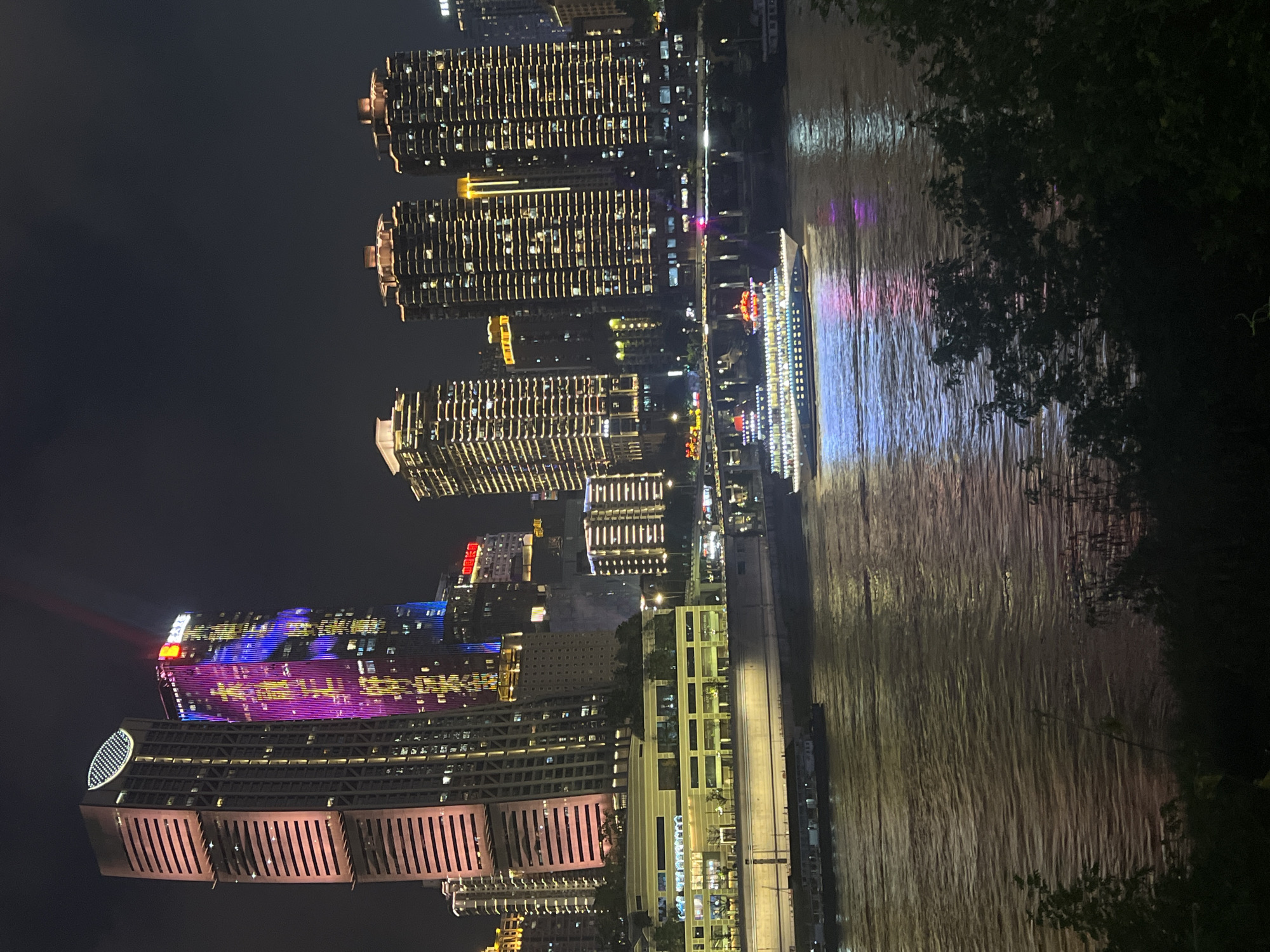 This image captures a nighttime scene along the Jialing River in Jiangbei Cheng Street, Chongqing, China. The waterfront is dominated by a cityscape of illuminated skyscrapers, with the most prominent being a tall, rectangular building on the left with horizontal light bands and a rounded, oval light fixture at its peak. To its right, another skyscraper is vertically illuminated with vibrant purple and yellow lights, with a red beam of light extending upwards from its roof.
Further down the river, several other modern buildings with rhythmic, horizontal lighting are visible. A large, multi-deck passenger boat, brightly lit with white and red lights, is cruising on the dark, rippling waters of the river, casting colorful reflections of its lights onto the water's surface. The foreground of the image shows the dark silhouettes of trees and foliage, suggesting the viewer is positioned on a bank or elevated viewpoint overlooking the river and the illuminated city. The overall atmosphere is one of urban vibrancy and nighttime spectacle.