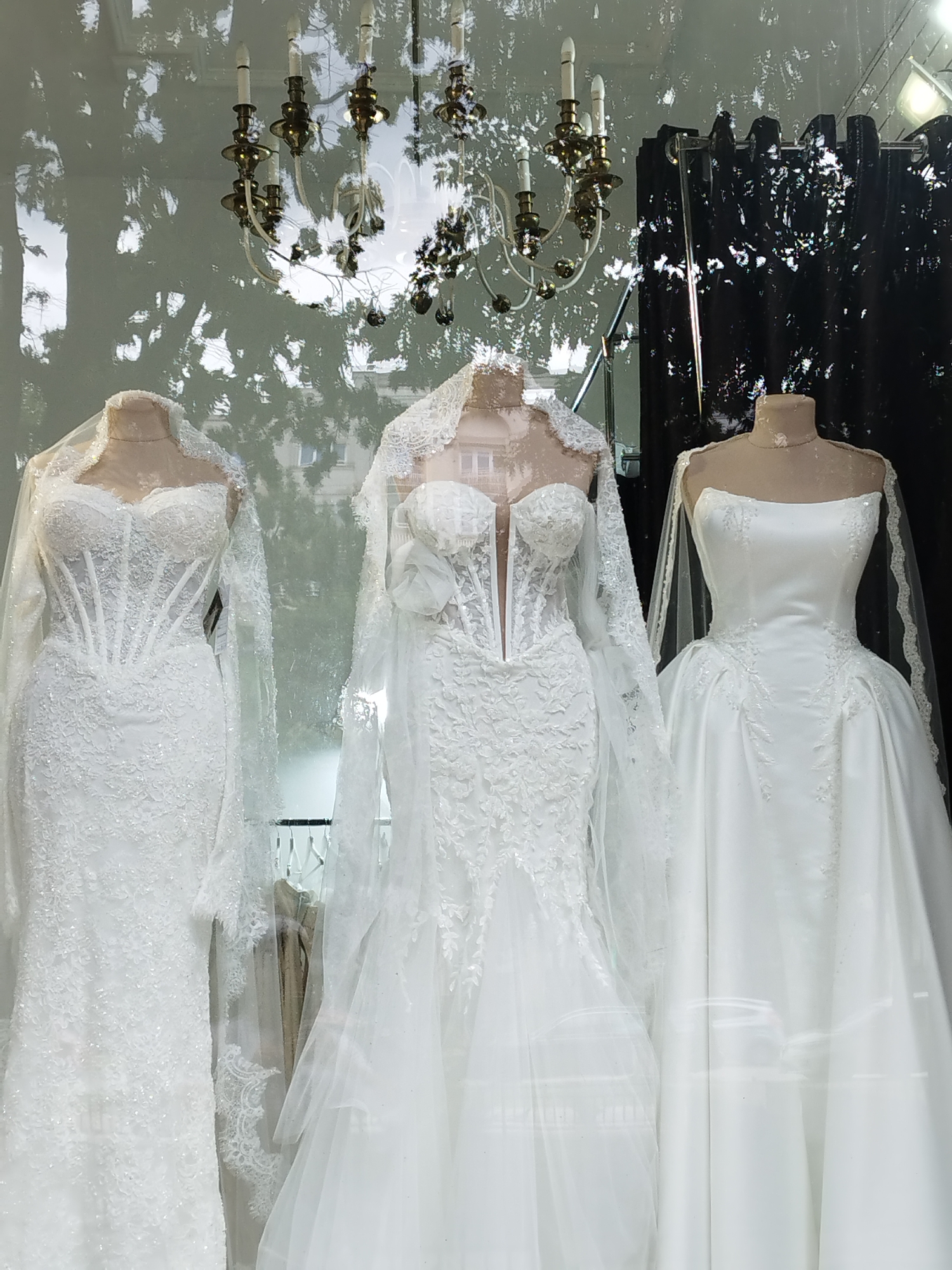 The image shows a storefront window displaying three white wedding dresses on mannequins. The dresses are elaborate, with lace and intricate detailing. A chandelier is visible in the reflection of the window, along with the silhouettes of trees and buildings. The scene is likely from a bridal shop located in Bucharest, Romania. The lighting is bright, suggesting it's daytime. The mannequins are positioned closely together, filling the frame of the window. The overall impression is one of elegance and sophistication, typical of a high-end bridal boutique.