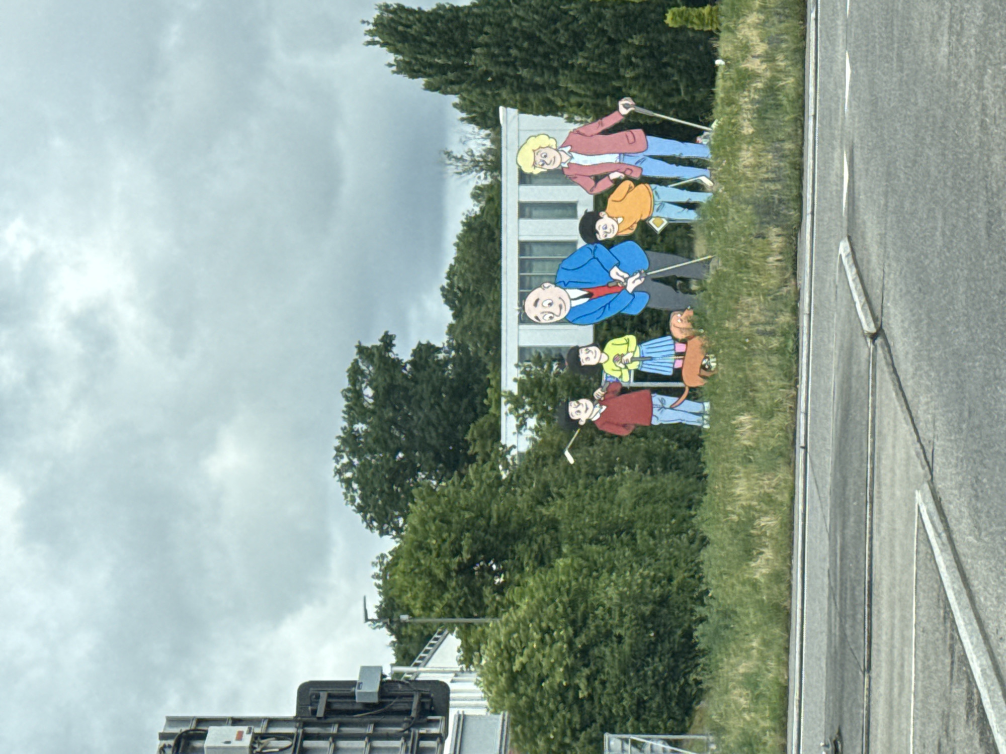 The image, taken in Zurzach, Switzerland, presents a vibrant roadside display featuring several cartoon-style cutouts of people playing golf. The scene is set on a grassy verge adjacent to a paved road, with lush green trees and a modern building in the background.
The cutouts portray what appears to be a family enjoying a golf outing. From left to right (when oriented correctly): an adult woman with blonde hair, wearing a red jacket and blue pants, holds a golf club. Beside her, a young boy with black hair in an orange shirt and blue pants looks on. A central figure is a bald adult man in a blue jacket, white shirt, and red tie, also holding a golf club. Further right, a girl with black hair, dressed in a yellow-green top and a blue-and-white striped skirt, poses with a golf club, accompanied by a small, reddish-brown animal. Another smaller individual in a red-brown top and blue pants holds a club, completing the golfing ensemble. All characters exhibit cheerful, smiling expressions.
The sky is overcast with light grey clouds, indicating a cloudy daytime. Behind the figures, verdant trees and bushes create a natural backdrop, and a contemporary, light-colored building with large windows is partially visible. The right side of the image features a multi-lane paved road with clear white markings. On the far left, parts of a large vehicle, possibly a truck or trailer, and some industrial equipment can be seen. This display appears to be a public art installation or an advertisement, likely related to a golf attraction in the vicinity. No discernible text is visible on the cutouts or surrounding objects.