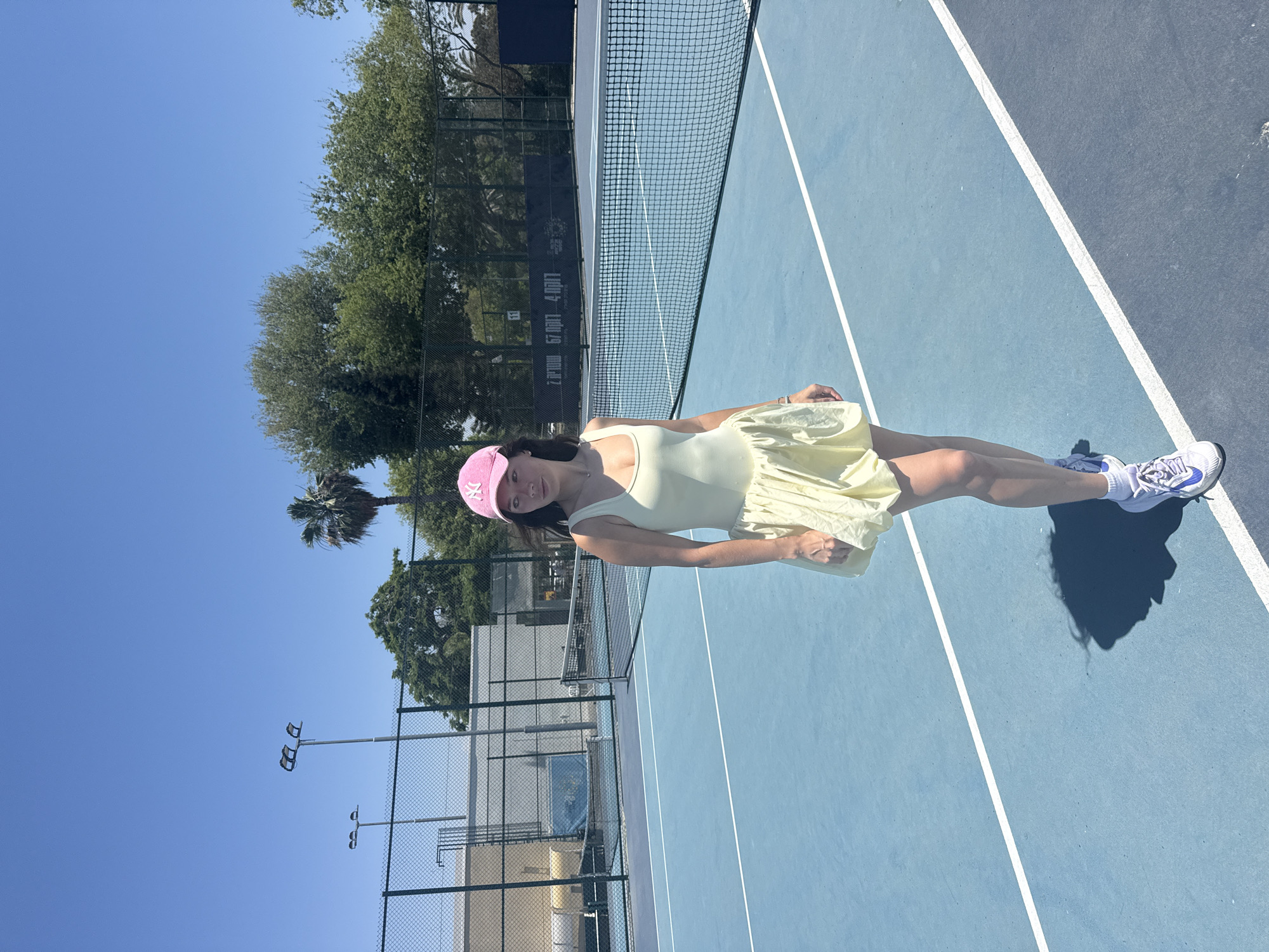 The image shows a woman in a light yellow outfit and a pink baseball cap standing on a tennis court. She wears white sneakers with blue accents. The tennis court has a blue surface with white lines and a black net. In the background, there are trees, a palm tree, and a fence surrounding the court. There are lights positioned above the fence. The sky is clear and blue. This image could be located in the city of Tel-Aviv, Israel, which is a possible location because Tel-Aviv has many tennis courts.