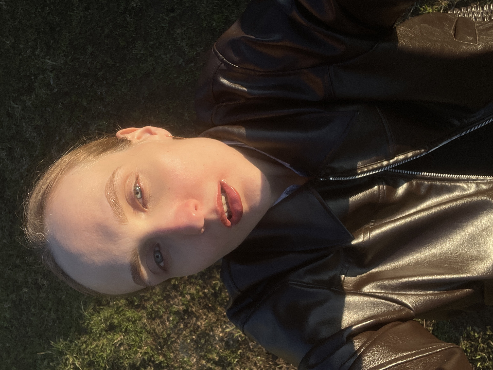 A young individual, likely female, is featured in a close-up portrait, lying on the ground outdoors and looking upwards towards the camera. She has fair skin, light-colored hair, and striking blue eyes. Her lips are slightly parted and appear glossy, suggesting lip gloss. She is wearing a dark brown or black leather jacket with a prominent silver zipper visible on the front.
The setting appears to be a natural outdoor environment, with green grass or moss forming the background beneath and around her. This aligns with the provided location context of Sovetsko-Nikol'skoe Lesnichestvo, Russia, implying a forest or wooded area.
The image is captured under strong, warm sunlight, casting distinct highlights on her face, particularly on her forehead and nose, and on the textured surface of her leather jacket. This lighting suggests either early morning or late afternoon. The overall angle is from below, with her head slightly tilted, giving the composition a dynamic feel. There is no visible text in the media file.