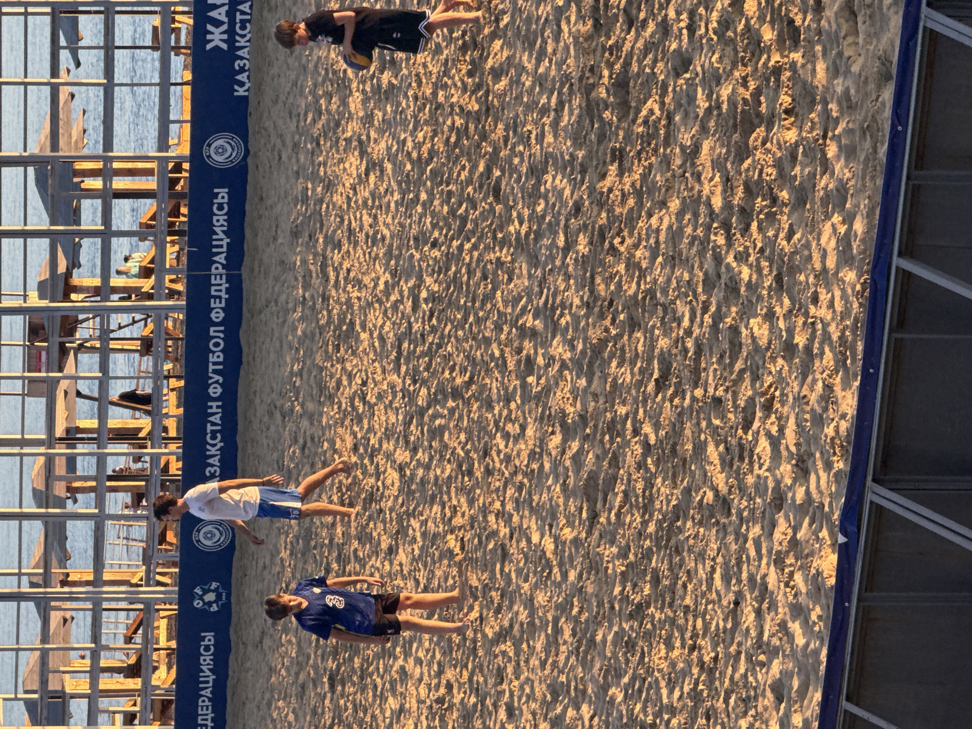 The image shows a beach volleyball match in progress on a sandy court, bathed in the warm glow of late afternoon sun. Three young men are visible on the court. One, wearing a black t-shirt and shorts, is in the process of hitting a volleyball. Another, in a white t-shirt and blue shorts, appears to be observing the game. The third, dressed in a blue jersey with the number "3" on it and black shorts, is also on the court.
Behind the players, a blue banner with white lettering runs along the edge of the court. The text on the banner is in Russian and appears to be related to the Kazakhstan Football Federation. Visible words include "ЗАҚ" and "ФЕДЕРАЦИЯСЫ". The setting is a beach, with the calm blue water of a lake or sea visible in the background, bordered by wooden structures. The sand is textured, with footprints and mounds indicating recent activity. The sunlight casts long shadows, confirming it is late in the day. The overall atmosphere suggests a friendly or competitive sports event.