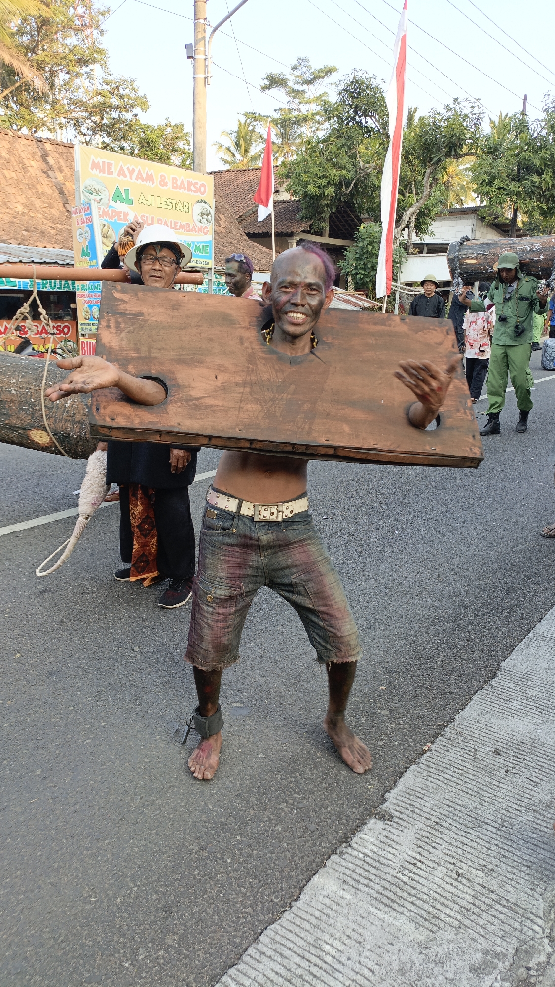 In Krajan Lor, Indonesia, a man with a dark complexion and purple-dyed hair is participating in a public event. He is wearing a wooden stock device around his neck and torso, with his arms protruding through the openings. He is also barefoot and has an ankle shackle on his left leg. He is wearing distressed denim shorts and a white studded belt.
The man is smiling broadly and gesturing with his right hand. Behind him, other people are visible, including a man in a white hard hat and glasses, and another man with dark face paint. A large wooden log is also present, attached to the stock device. In the background, there are buildings, trees, and Indonesian flags displayed. The event appears to be taking place outdoors on a paved road, possibly during a parade or festival.