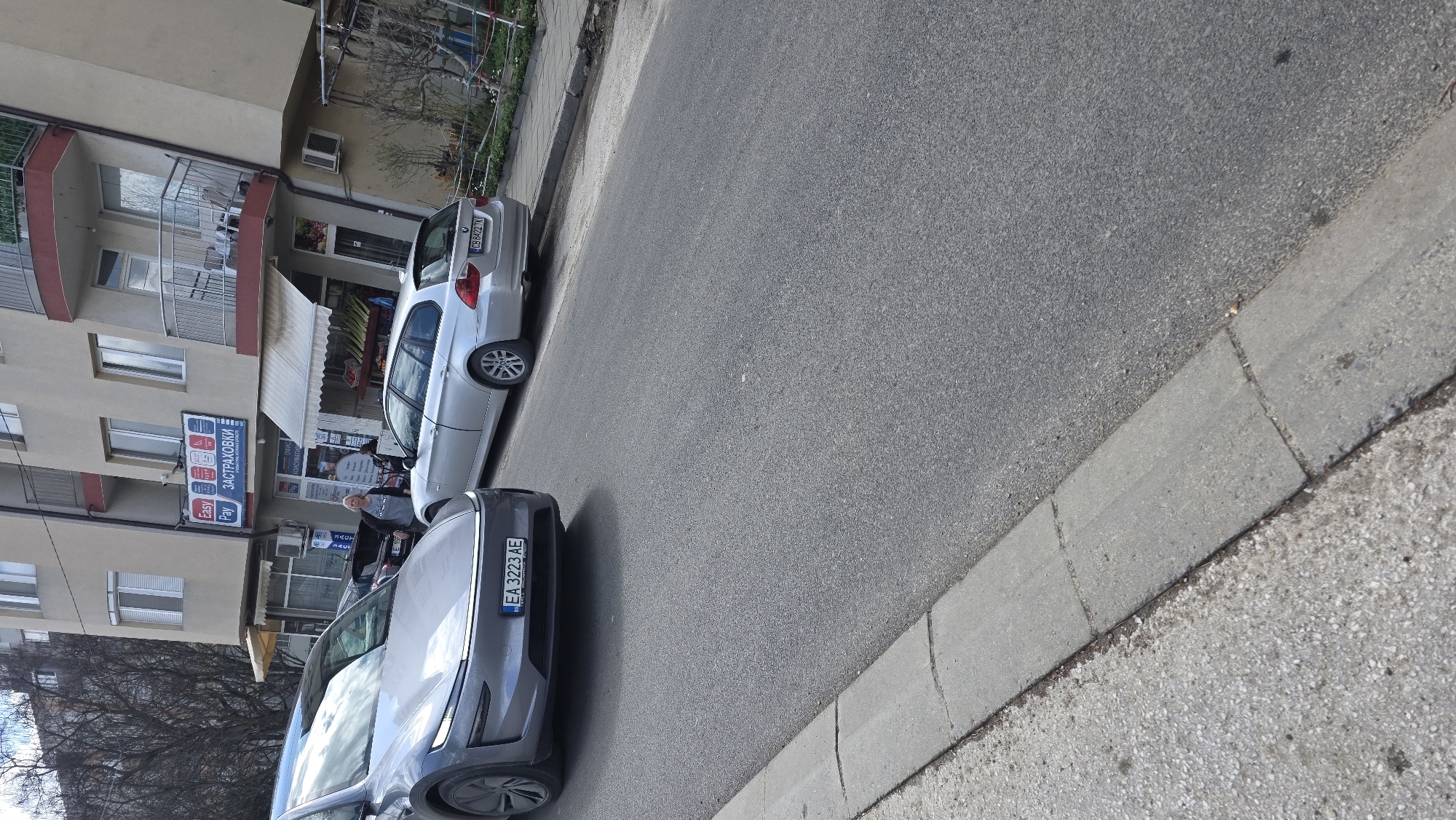 The image depicts a street scene in Blagoevgrad, Bulgaria. Two cars are parked along the side of the road. The car in the foreground is grey, with license plate "EA 3223 AE". Behind it is a silver car, license plate "A08342nx". The cars are parked in front of a building with a shop on the ground floor. The building appears to have multiple floors. People are seen in front of the shop, and there are signs for "Easy Pay" and "ЗАСТРАХОВКИ" (Insurance) visible on the building. The street is paved with asphalt. A concrete curb is adjacent to the road.