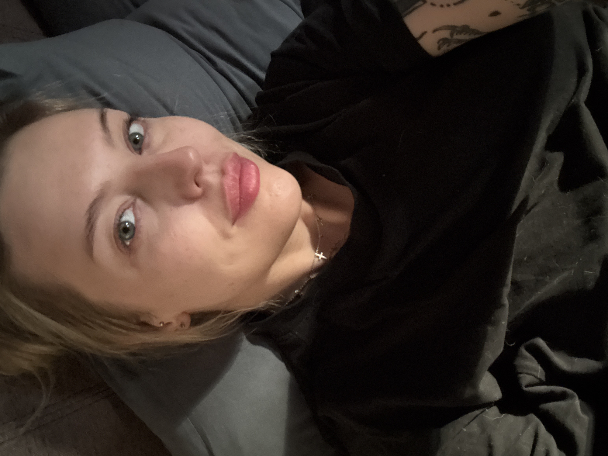 A woman is lying on a gray pillow and covered with a dark, possibly black, garment. She has fair skin, blonde hair, and light-colored eyes. A silver necklace with a cross pendant is visible around her neck. She appears to have a piercing in her nose. There are tattoos on her arm. The environment appears to be indoors, possibly a bedroom or living room, based on the presence of bedding. No information suggests the location is in Krasnoyarsk, Russia.
