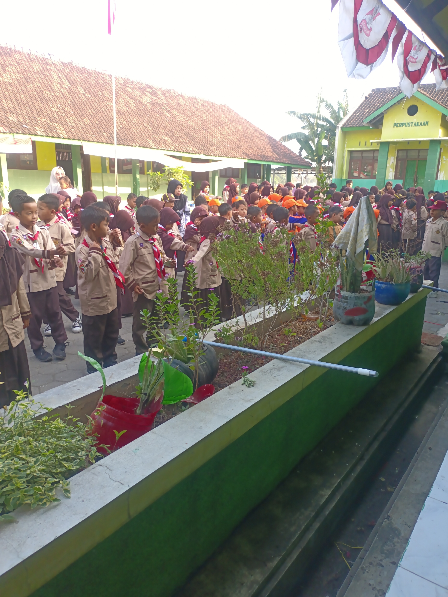 In Sarimulyo, Indonesia, a group of students, likely in uniform, are gathered outdoors in front of a building. The students are wearing tan uniforms with red and white neckerchiefs, suggesting they are part of a scout troop or similar organization. Many are wearing headscarves, indicating a mixed-gender group or that the female students are wearing them as part of their uniform. Some students are wearing orange hats.
The students are standing in a line, facing towards the right of the image. In the foreground, there is a raised planter box filled with plants and decorated with colorful recycled materials, including a red plastic container and a blue pot. A white cloth draped over a plant adds an unusual element. A metal pipe runs along the top edge of the planter box.
Behind the students, two buildings are visible. The building on the left has a tiled, sloping roof and a yellow wall. The building on the right is painted green and yellow, with "PERPUSTAKAAN" (Library) written above a window. Flags with red and white designs are hanging from a pole above the buildings. The environment appears to be sunny, with shadows cast on the ground. The overall scene suggests a school event or assembly.