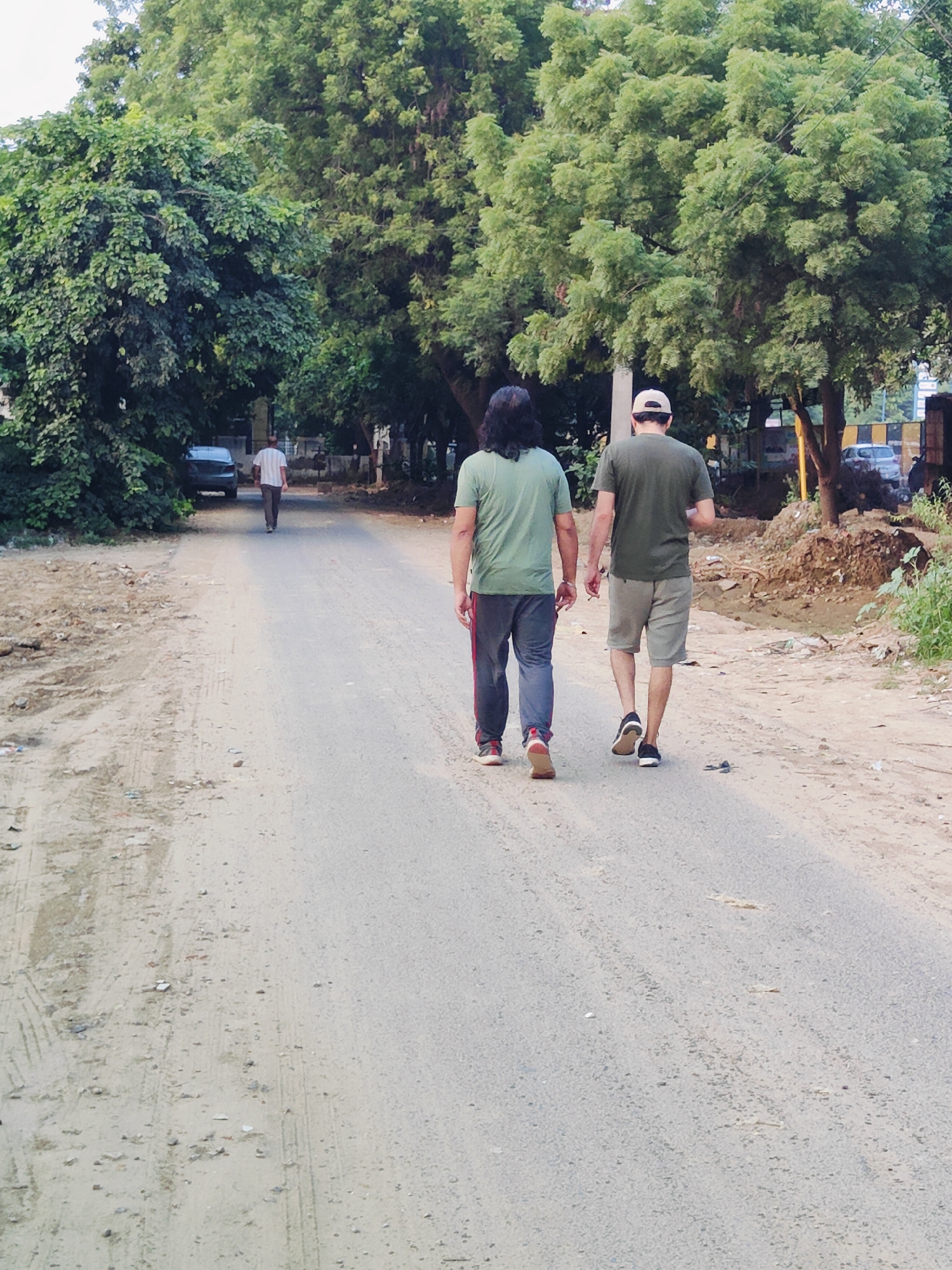 A long shot captures two men walking away from the camera on a dirt and asphalt road, bordered by lush green trees and vegetation, suggesting an outdoor, semi-urban environment within Gurgaon, India. The man in the foreground on the left wears a green t-shirt and dark pants with red stripes, while the man to his right wears a green t-shirt and light-colored shorts. A third, distant figure in a white and striped shirt walks further down the road, also away from the camera. Cars are visible parked or driving in the background. The road surface is uneven, with patches of dirt, gravel, and what appears to be worn asphalt. The lighting suggests it is daytime, possibly late afternoon, with diffused sunlight filtering through the dense foliage. The overall scene depicts a casual stroll in a somewhat unkempt, natural setting.