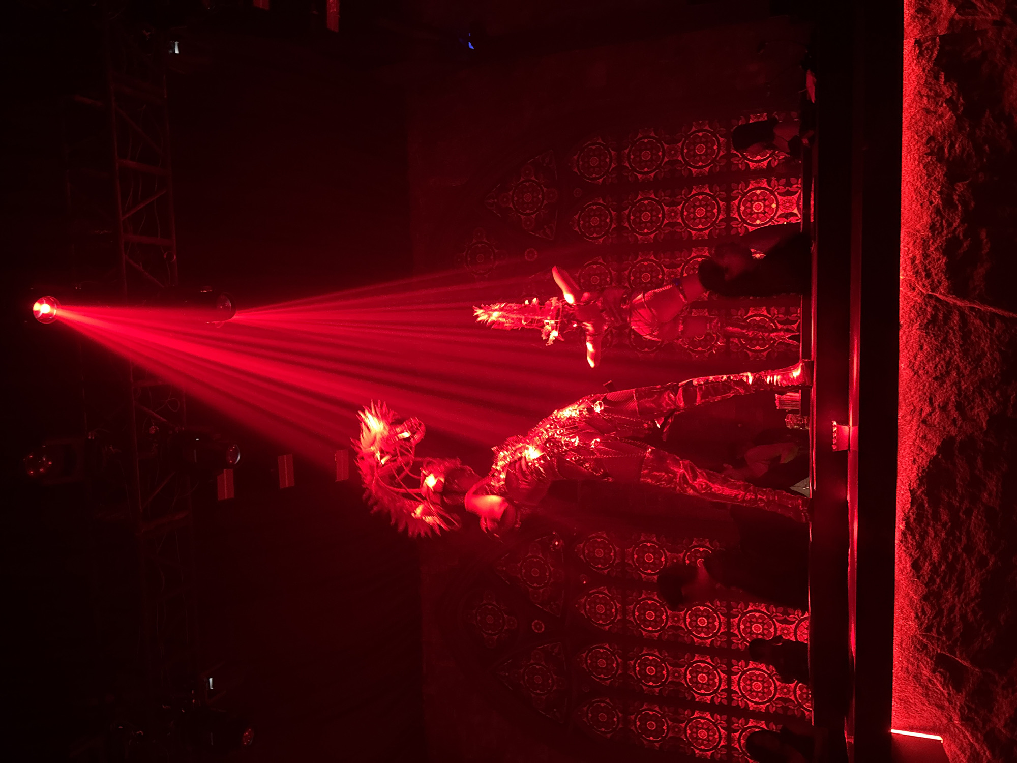 In a dimly lit, red-hued nightclub in North Kuta, Indonesia, two performers in elaborate, shimmering costumes command the stage under a dramatic red spotlight. The performer on the left, positioned slightly further back, wears a fitted, metallic outfit adorned with intricate patterns and a large, spiky headpiece resembling flames or a mythical creature. They are captured in a dynamic pose, leaning forward with one arm extended. To the right and slightly forward, another performer, similarly dressed in a metallic costume and a distinctive headpiece, stands in a more upright posture. The backdrop features a repetitive, circular, mandala-like pattern in a darker shade of red, creating a visually textured wall. In the foreground, a bar area is visible with silhouetted figures of patrons seated and standing, their faces obscured by shadow. The overall atmosphere is one of vibrant nightlife and performance, amplified by the intense red lighting and the striking costumes.