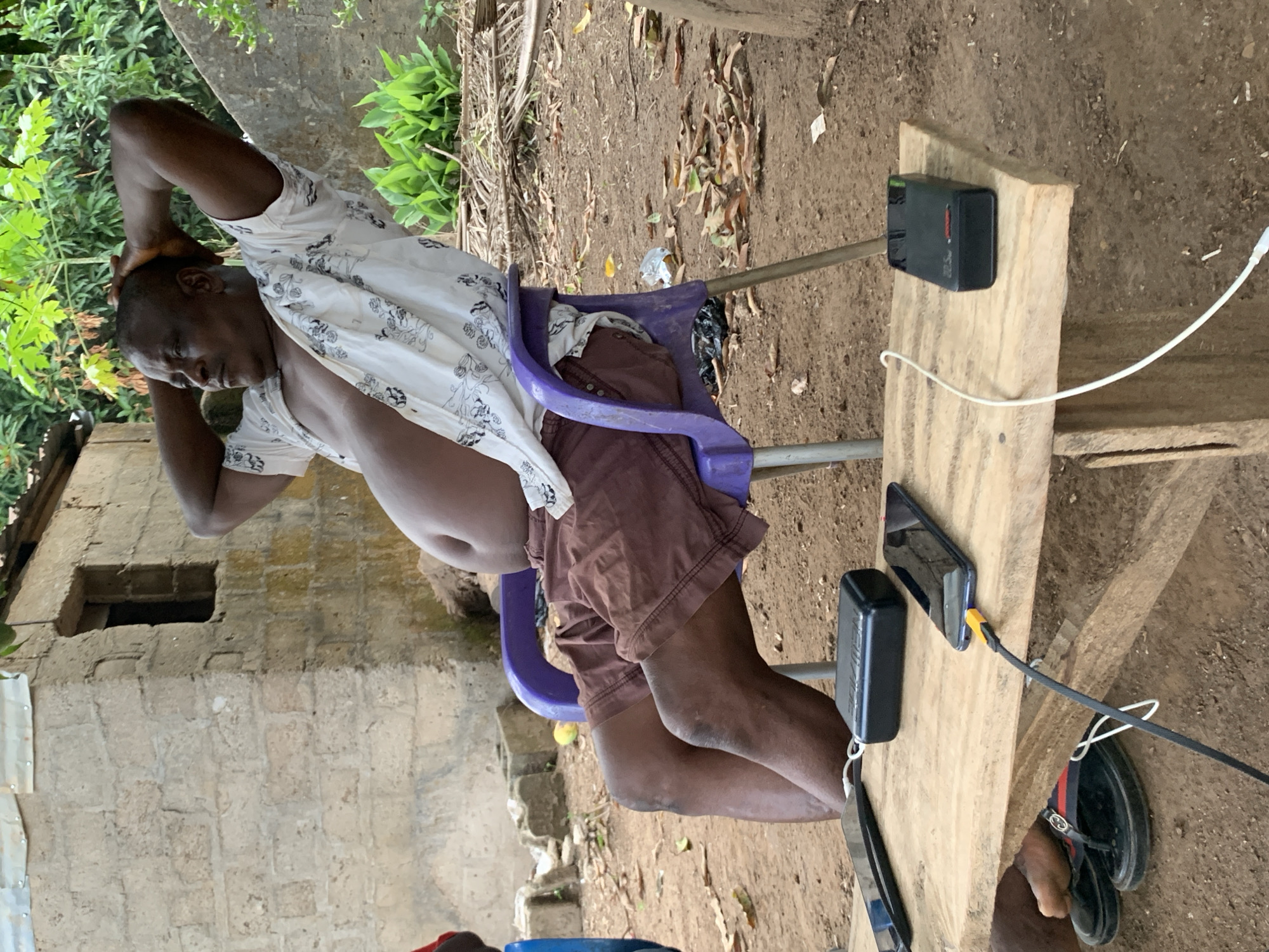 A man is sitting on a purple plastic chair outdoors in Akwanga, Nigeria. He has his hands behind his head and is wearing a white patterned shirt and brown shorts. He is positioned next to a wooden table that holds a smartphone, a black rectangular device connected by wires, and a pair of black sandals. The environment includes dirt ground, vegetation, and a partially visible brick building.