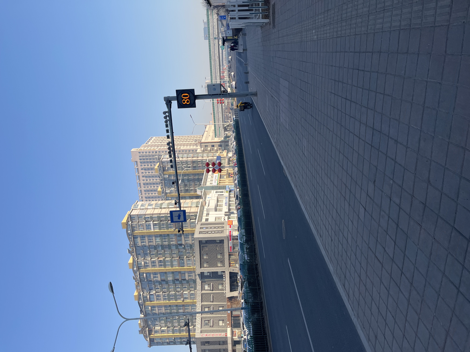The image depicts a street scene in Changchun City, China. The street is divided by a median with a green barrier. A digital speed limit sign reads "80." Buildings line one side of the street, while a wide, tiled sidewalk runs along the other. A yellow motorized scooter is visible on the street. The sky is clear and blue.