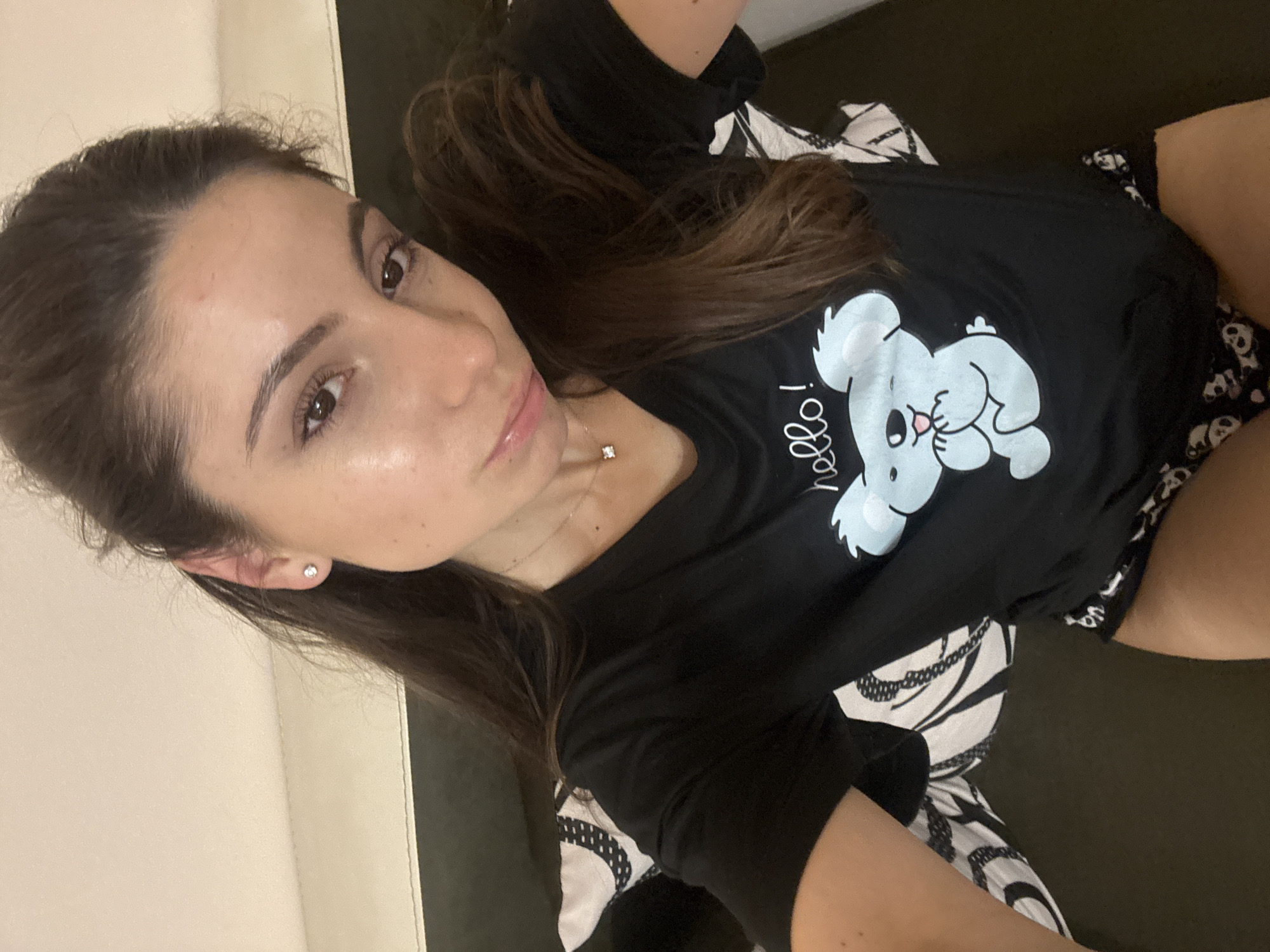 A young woman with long, dark brown hair is lying down on a bed. She is wearing a black t-shirt with a cartoon koala graphic that says "hello!" and black underwear with a panda print. She has a small diamond necklace on. The background appears to be a bed with white and black patterned sheets. The lighting suggests an indoor setting, possibly a bedroom. No specific location in Varna, Bulgaria is indicated.
