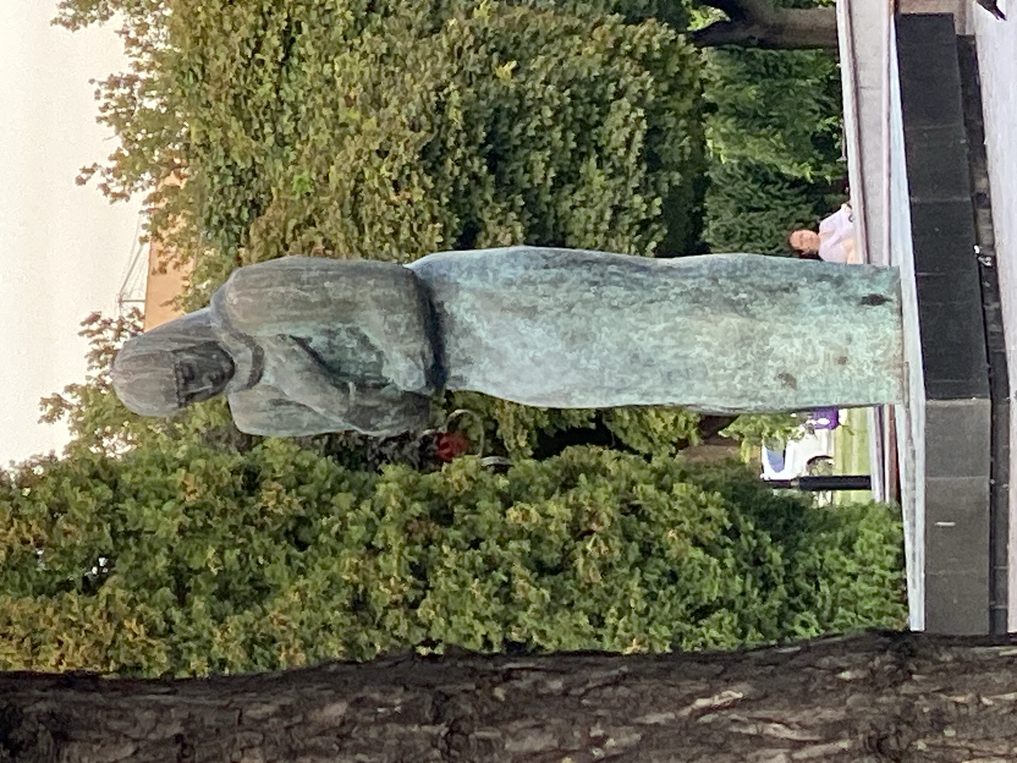 A bronze statue of a somber-looking woman in a hooded robe stands in a park in Chișinău, Moldova. The statue is placed on a stone pedestal. Behind the statue, there are lush green trees and bushes. In the background, a white car is visible. To the right of the statue, a woman in a light-colored shirt is sitting on a bench. The sky is overcast.