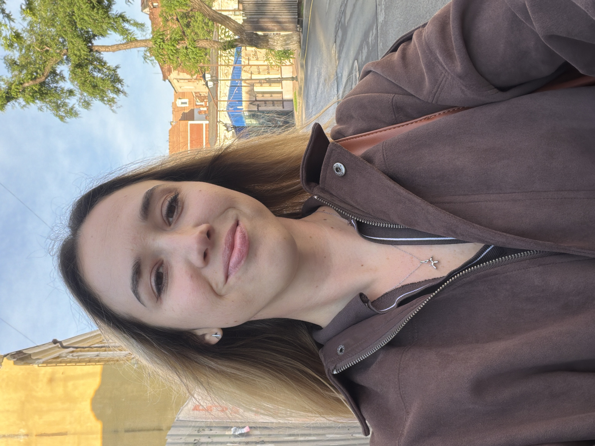 A woman is taking a selfie. The woman has shoulder-length brown hair with blonde highlights and light skin. She is wearing a brown jacket over a dark-colored top. A silver necklace with a pendant is visible.
The background shows an urban setting. Buildings are visible with some possibly under renovation, with blue tarps visible. A tree is to the side. The lighting suggests it is daytime. Based on architectural elements and the visible street, the location is likely within the city of Rostov-on-Don, Russia.
