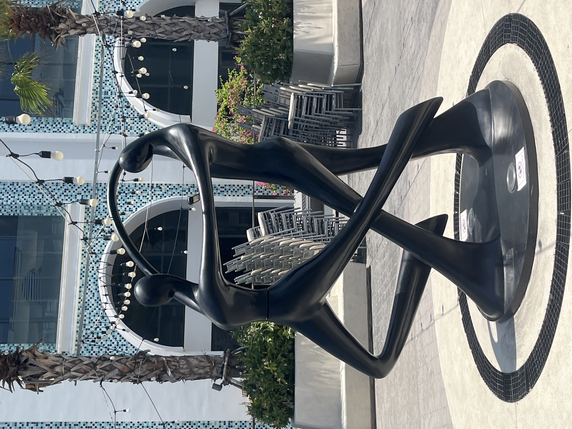 This image shows a black abstract sculpture of two figures dancing in a circular paved area. The sculpture is located in Nha Trang, Vietnam, in front of a building with white and blue tiled accents and arched windows. String lights with white bulbs are visible hanging in the background. Palm trees and green bushes are also present. Stacked chairs are visible behind the sculpture. The overall scene suggests an outdoor public space, possibly a plaza or walkway, near a commercial establishment.