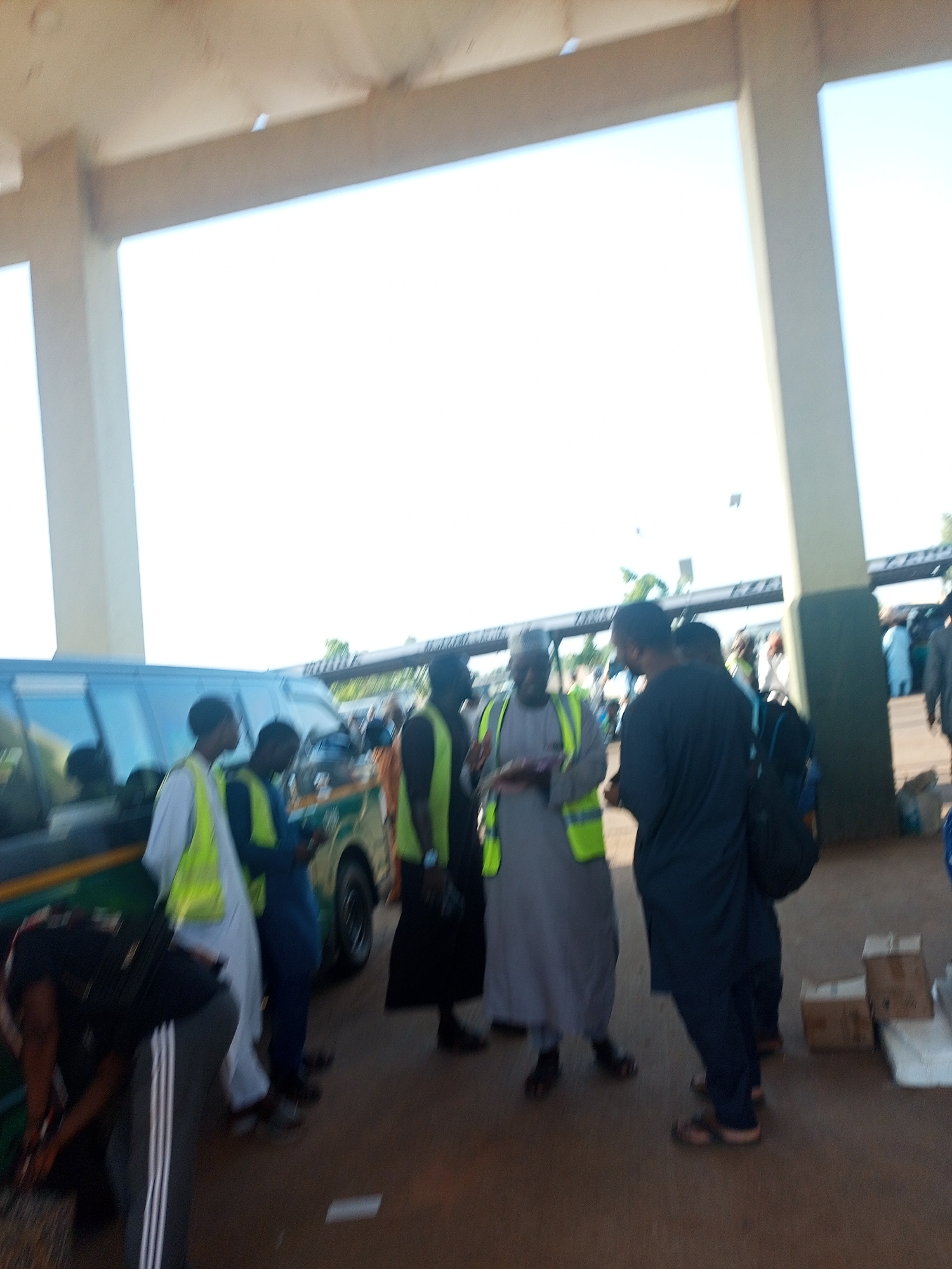 The image shows a group of people standing near a bus at what appears to be a bus station or transport hub. Several individuals are wearing yellow safety vests. One man is bending near the front of the bus, potentially loading or unloading items. The setting suggests an urban environment during daylight.