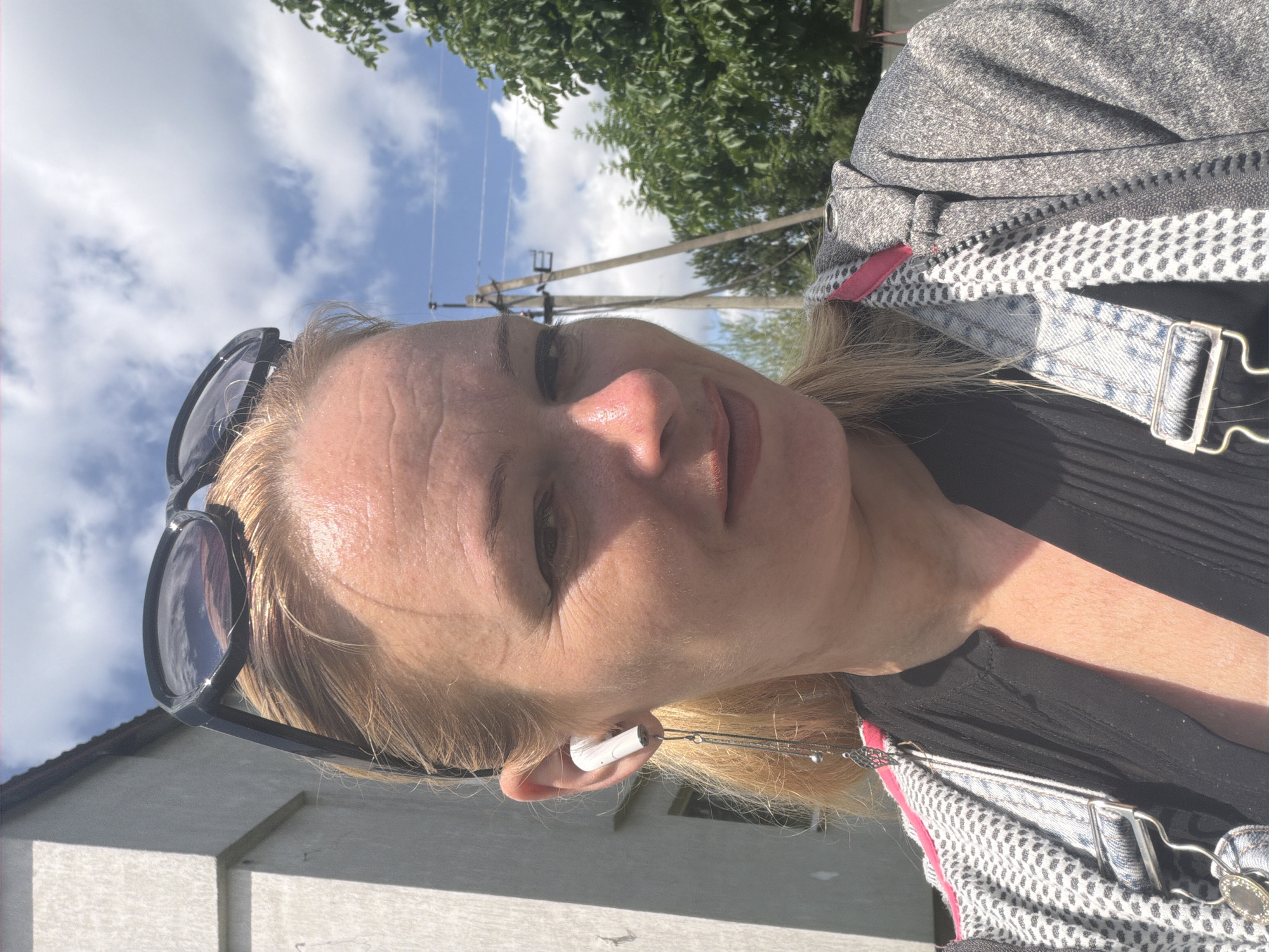 A woman with blonde hair and sunglasses perched on her head takes a selfie outdoors. She is wearing a black shirt and a grey zippered jacket with a pink lining. White earbuds are in her ears, with one dangling a long, decorative charm. The background shows a building, trees, a utility pole with wires, and a cloudy blue sky.