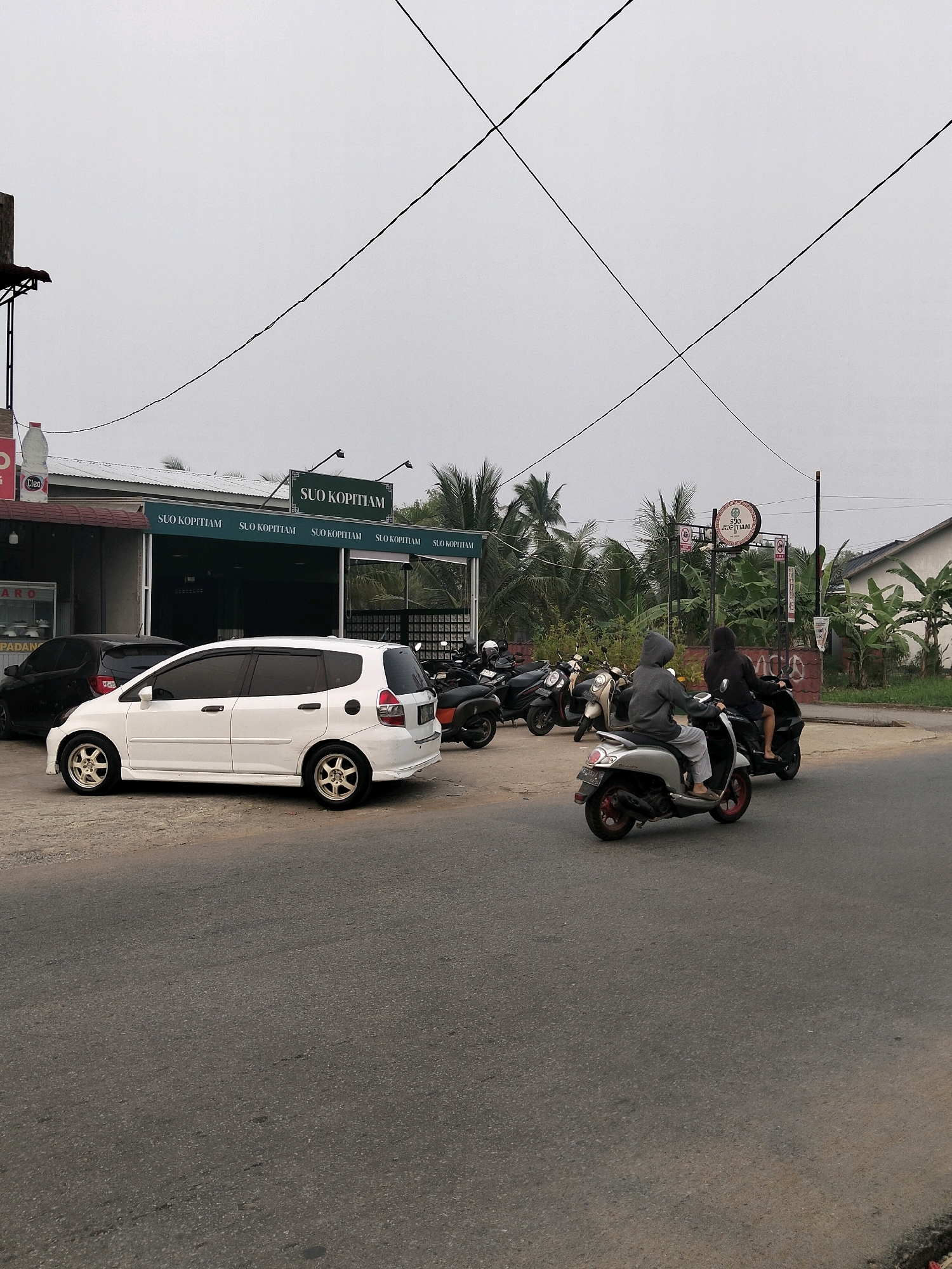 The image depicts an overcast day in a semi-urban area, likely Arang Limbung, Indonesia. In the foreground, a grey asphalt road runs diagonally across the frame. To the left, a white hatchback car is parked on the side of the road. Behind the car is a dark-colored car, also parked. Further left, a building with a green awning and multiple signs displaying "SUO KOPITIAM" is visible. Above the building, electrical wires crisscross the sky. To the right of the white car, two people are riding a silver scooter, with the rider in front wearing a grey hooded jacket and the passenger behind wearing a black hooded jacket. They are traveling on the road, moving away from the viewer. To the right of the scooter riders, a black scooter is parked, and further back, a row of parked motorcycles is visible in front of the building. Behind the building, a dense line of palm trees and other tropical foliage creates a green backdrop. The sky is a uniform light grey, suggesting cloudy or hazy weather.