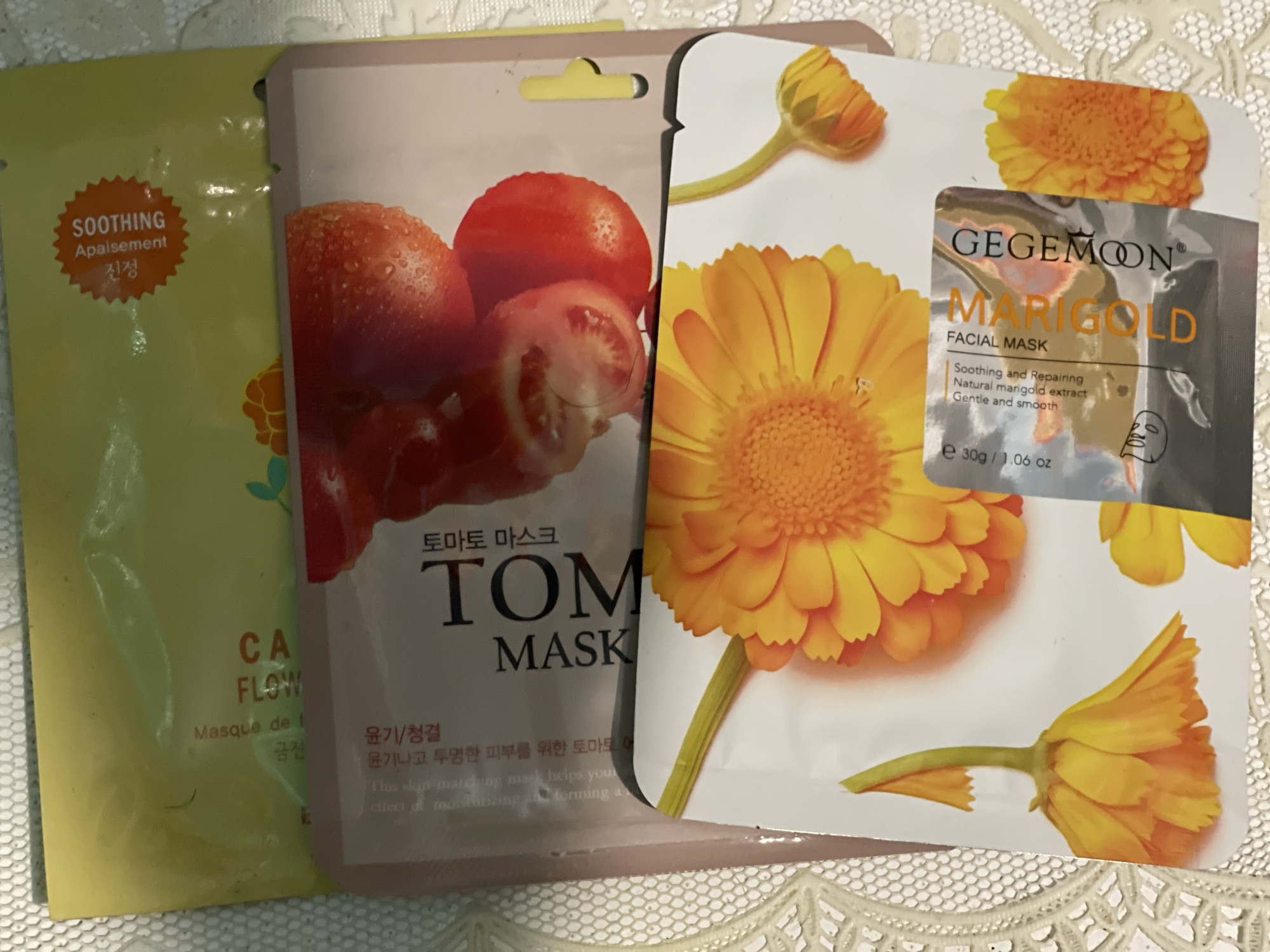 The image shows three facial masks laid out on a white lace-like surface. The subjects are the masks themselves, which are packaged in colorful wrappers.
The mask on the far left is yellow and features text in English and Korean, including "SOOTHING," "Apaisement," and "진정." Below this, there is text that appears to be "CA FLOW" and "Masque de f," along with some Korean characters. There is also a green floral graphic on the left side.
The middle mask has a pinkish-tan background and features a close-up image of several tomatoes, some of which are whole and others are cut. Text in Korean and English is visible, including "토마토 마스크" (Tomato Mask), "TOM MASK," and "윤기/청결" (Luster/Cleanliness). There are also descriptions in English, such as "This skin-matching mask helps you..." and "effect of moisturizing forming a...".
The mask on the right is white and features a large, vibrant orange marigold flower. Text in English is prominently displayed, including "GEGEMOON," "MARIGOLD FACIAL MASK," and "Soothing and Repairing Natural marigold extract Gentle and smooth." It also indicates the weight as "e 30g / 1.06 oz."
The scene is a close-up shot of these cosmetic products. There are no people visible, and the activity appears to be a static display of the items, possibly for product photography or review. The lighting suggests it might be indoor lighting, possibly natural light from a window, but it is difficult to determine the exact time of day or weather conditions. The white lace surface suggests a domestic setting or a craft-related background. The location context is given as "Средний, Russia." The overall impression is one of product presentation for skincare.