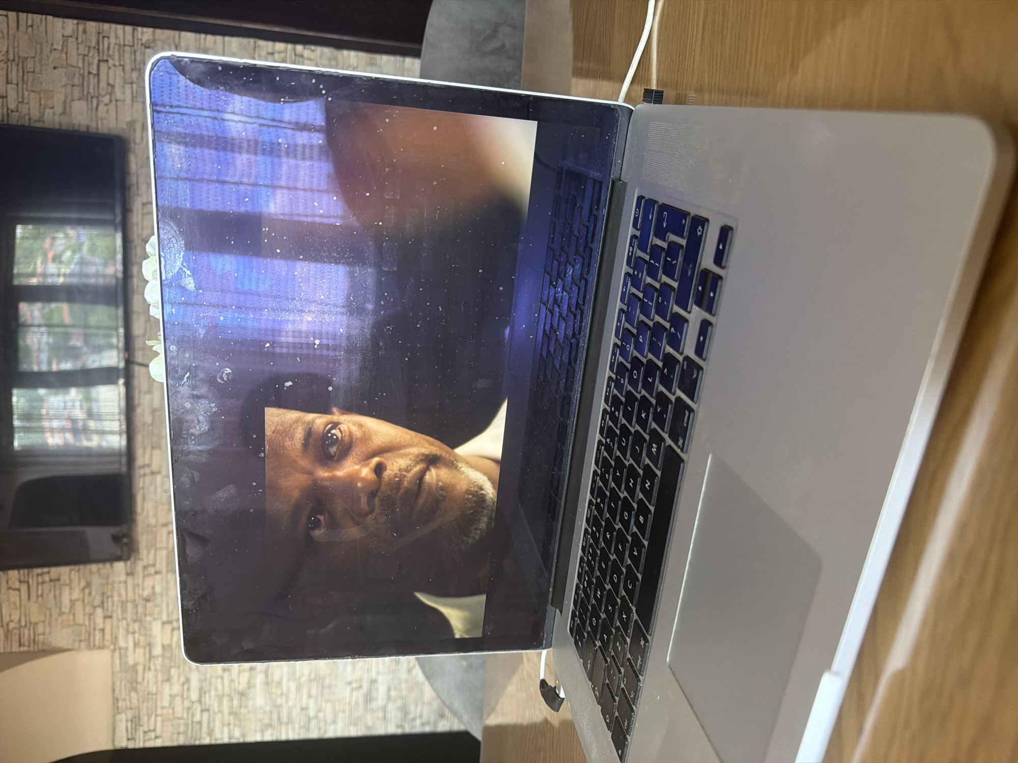 An overhead shot, rotated 90 degrees clockwise, captures a silver MacBook Pro laptop resting on a light-colored wooden surface in Shumen, Bulgaria. The laptop's screen is open and displays a close-up image or video of a man's face. The man has dark skin and a short beard, looking upwards and slightly to the right with a somewhat pensive or observant expression.
The laptop screen is noticeably dusty and dirty, covered in numerous small white specks. The top-left corner of the screen bezel shows signs of wear or damage, with a white, slightly jagged area. Below this, on the left side of the screen, there's a larger, irregular white smudge or possible damage to the screen protector.
A white charging cable is connected to the right side of the laptop, extending upwards out of frame. White earbuds are also plugged into the laptop's right side, near the charging port. The keyboard, visible on the lower half of the laptop, has black keys with white Latin characters (e.g., QWERTY layout) and a silver trackpad below.
In the background, on the left side of the image, a dark rectangular object, likely a television screen, reflects a window with horizontal blinds or curtains and what appears to be outdoor greenery. The wall behind this TV has a textured pattern resembling bricks or natural stone. The overall lighting suggests it is daytime, with natural light illuminating the scene.